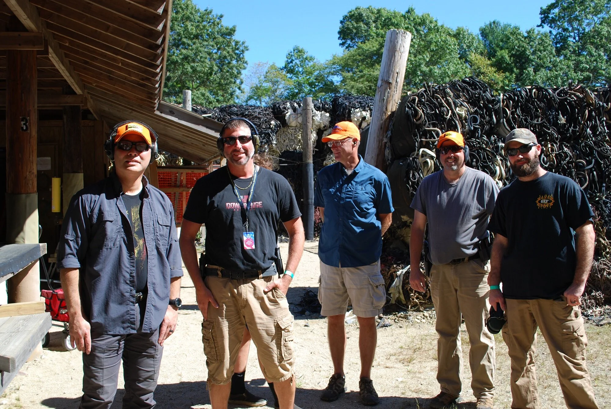 Five men standing outdoors on a sunny day, surrounded by trees and a large pile of black rubber in the background. They are dressed casually, some wearing sunglasses and hats.