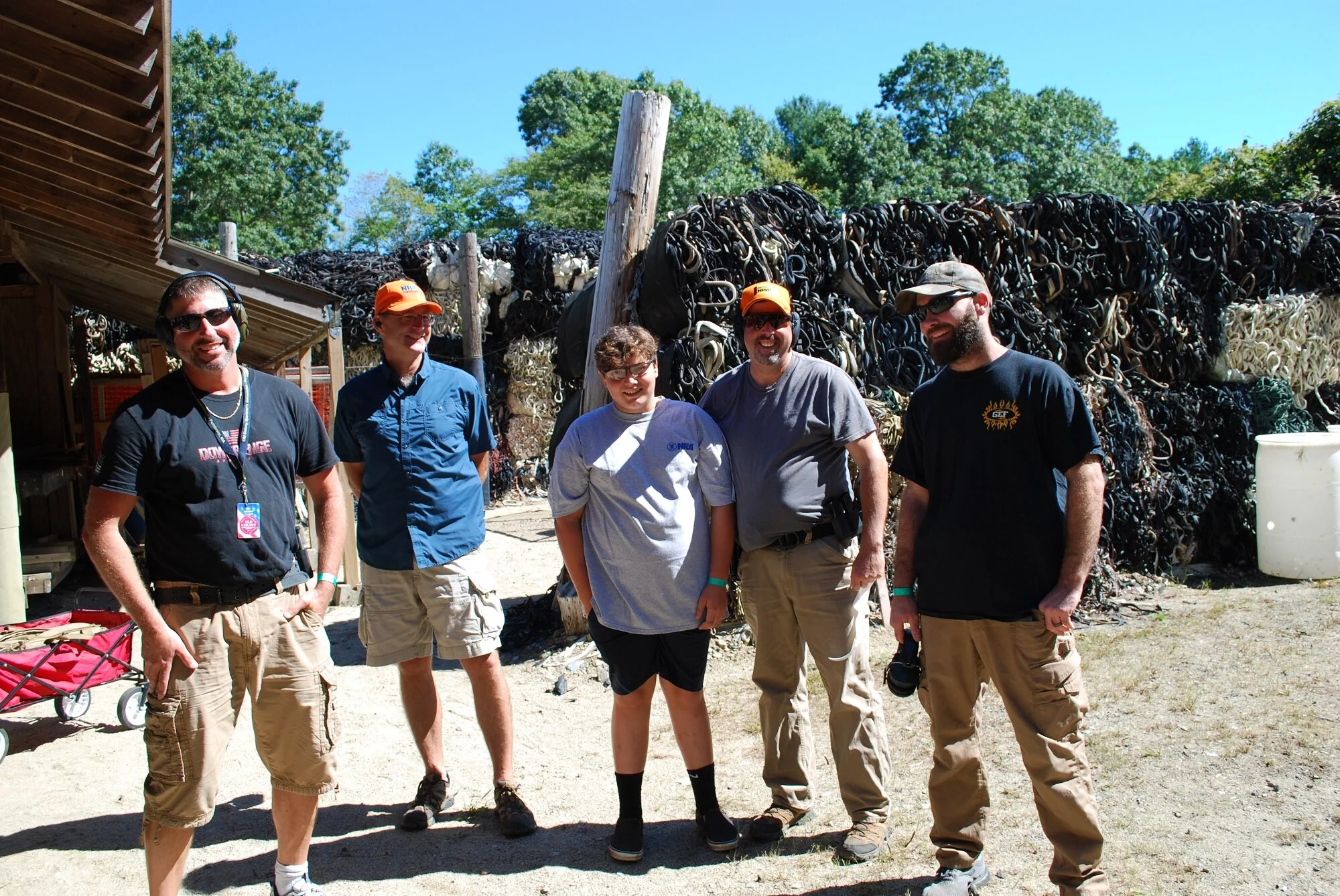Five people standing outdoors in front of a large pile of rubber or plastic tubing and a wooden structure, with green trees and blue sky in the background, smiling and posing for the photo.