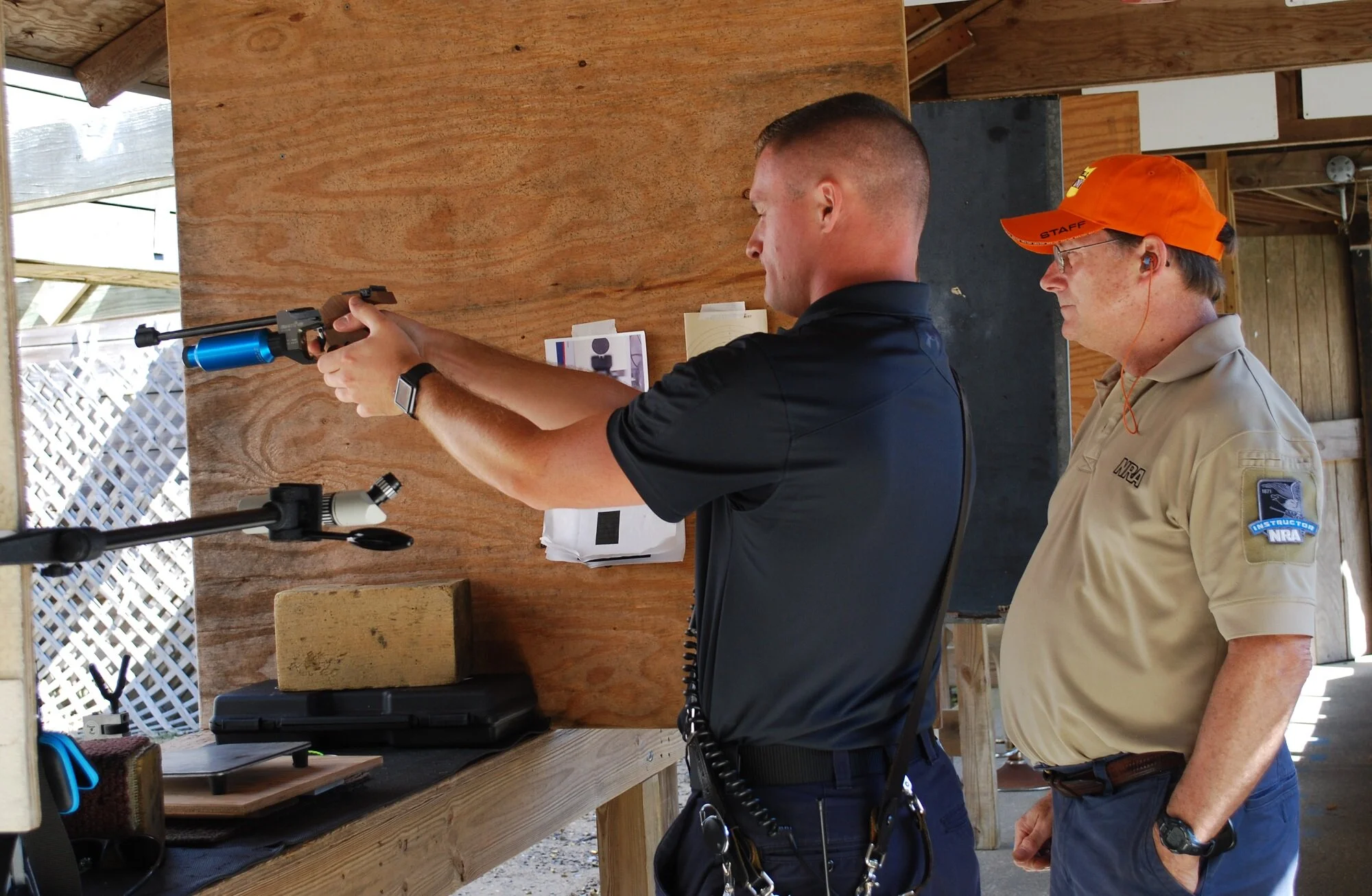 A man firing a rifle at a shooting range, with a staff member supervising, both standing in front of a wooden backstop, in an outdoor shooting range setting.