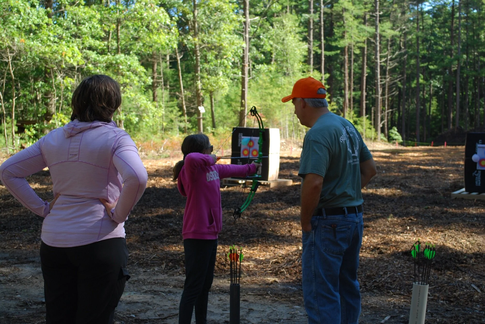 A girl in pink shooting an arrow at a target under supervision of two adults in an outdoor forest setting.