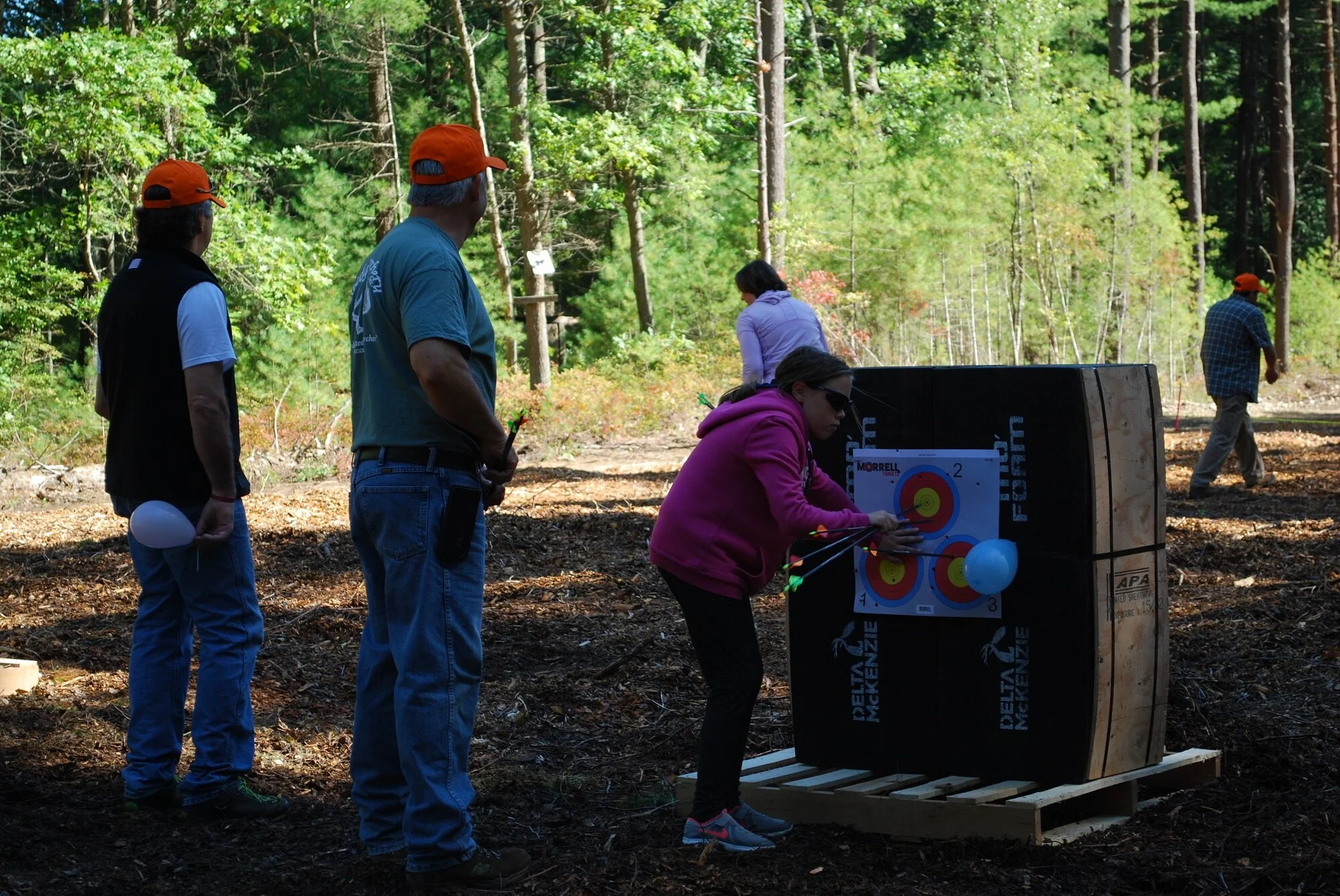 People participating in an outdoor archery activity in a wooded area; one person is aiming at a target, while others observe.