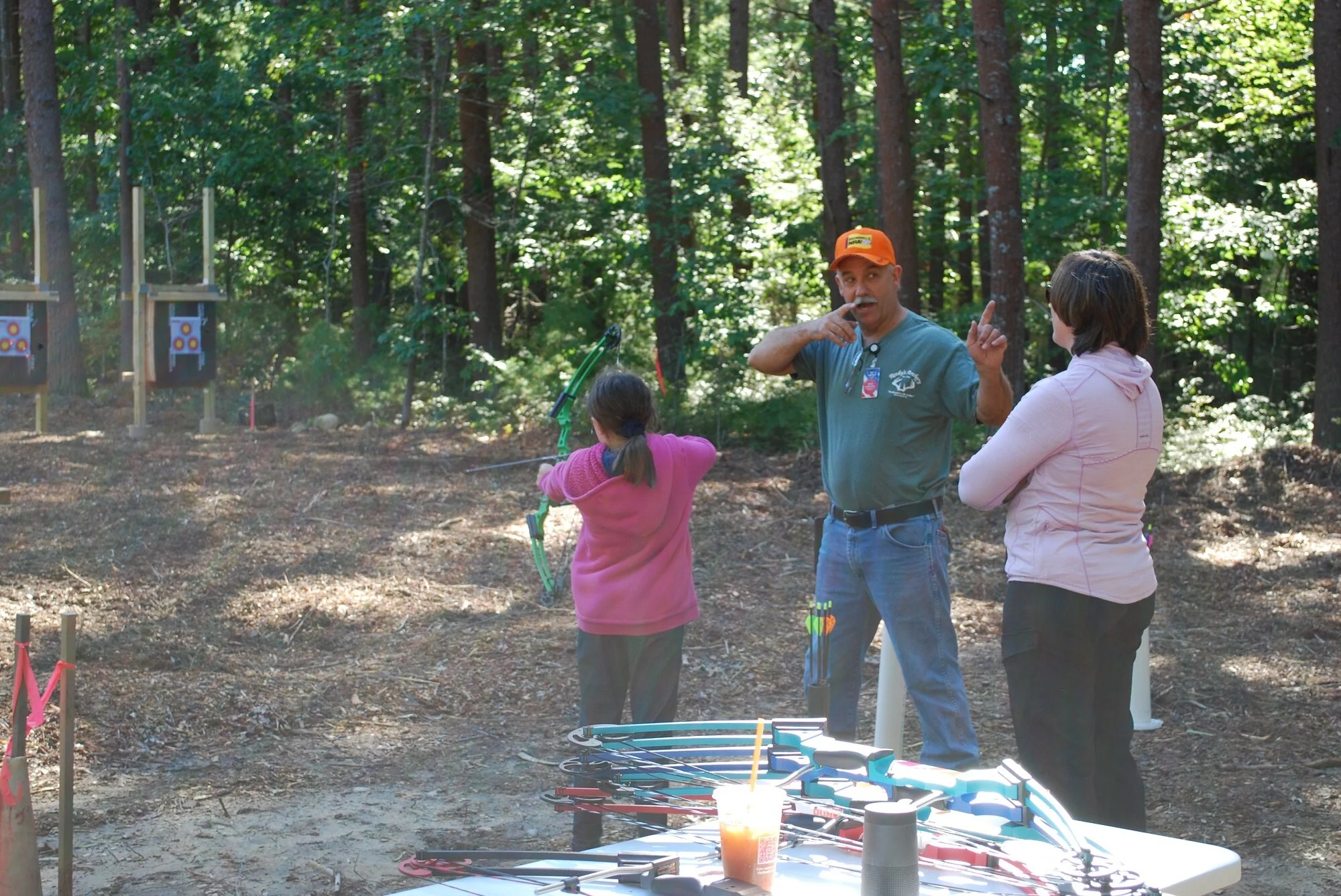 A girl practicing archery in a forested outdoor area with an instructor and a woman observing. There are targets on stands in the background and bows and drinks on a table in the foreground.