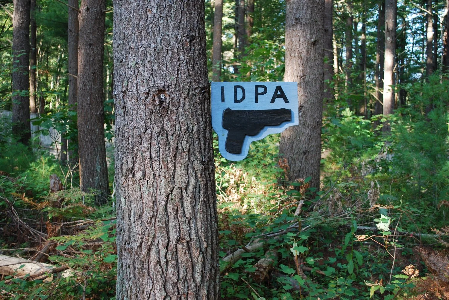 A sign attached to a tree in a forest with the letters 'IDPA' and a black silhouette of a handgun.