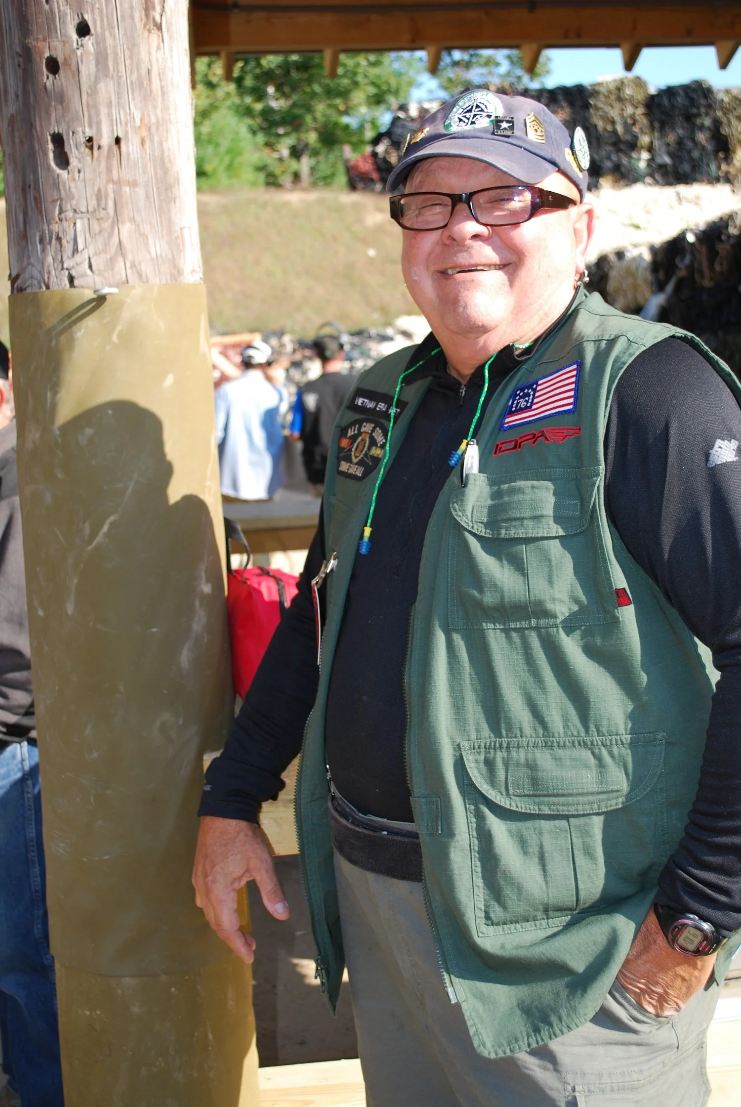A smiling man wearing glasses, a cap with patches, a green vest, a black shirt, and a wristwatch stands outdoors near a wooden pole. There are people and green trees in the background.