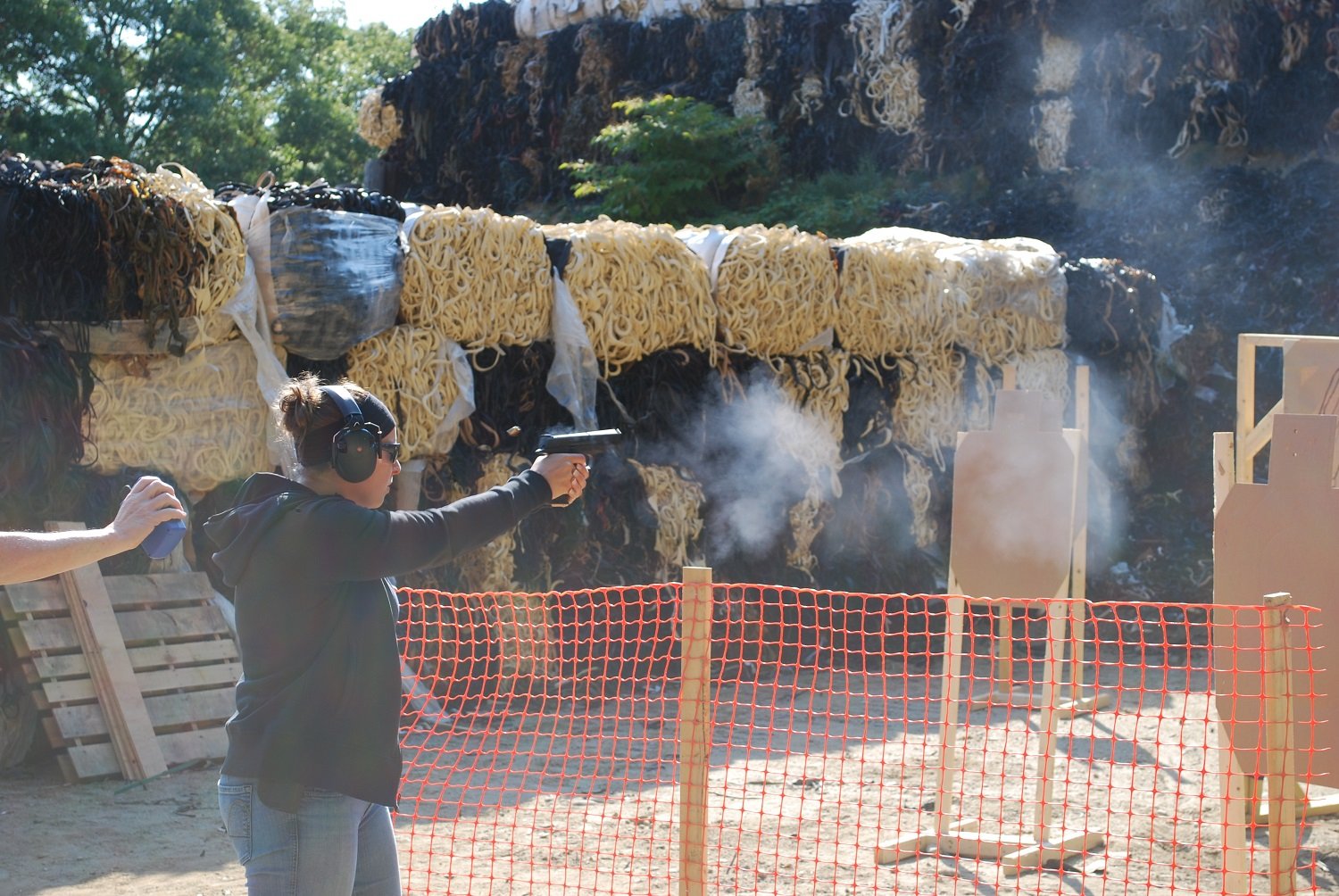 A woman at a shooting range is aiming a handgun, wearing safety headphones, with protective barriers and targets in the background.