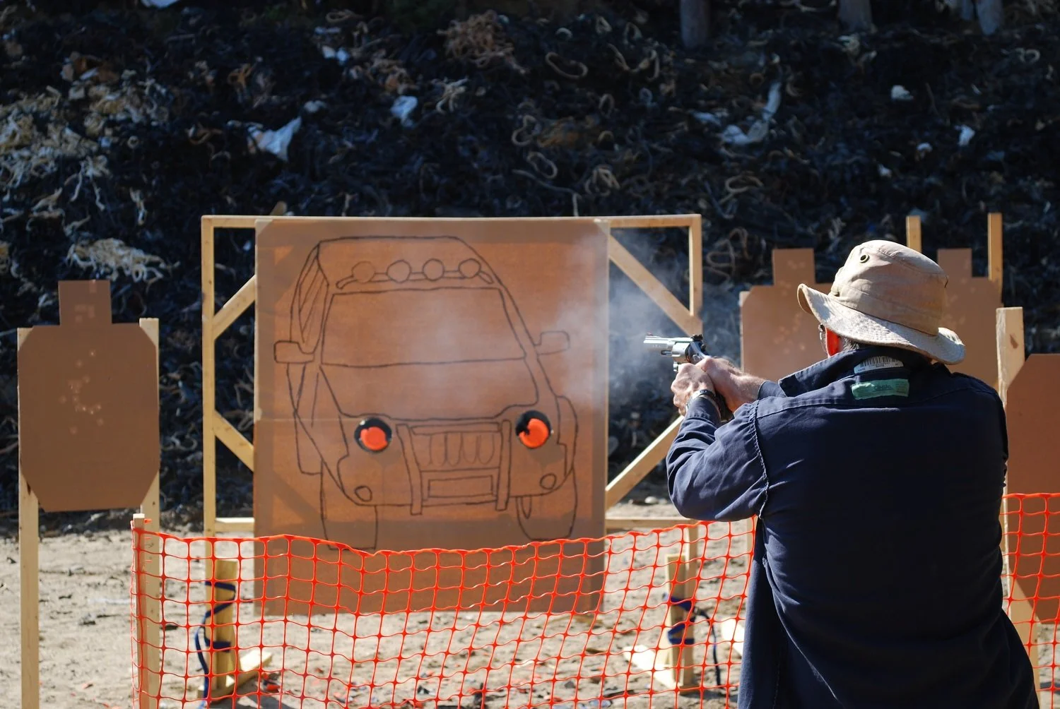 A person wearing a beige wide-brimmed hat and dark clothing shooting a handgun at a shooting range. They are aiming at a large cardboard target with a drawn outline of a vehicle. The background has a pile of discarded materials and a red plastic safe