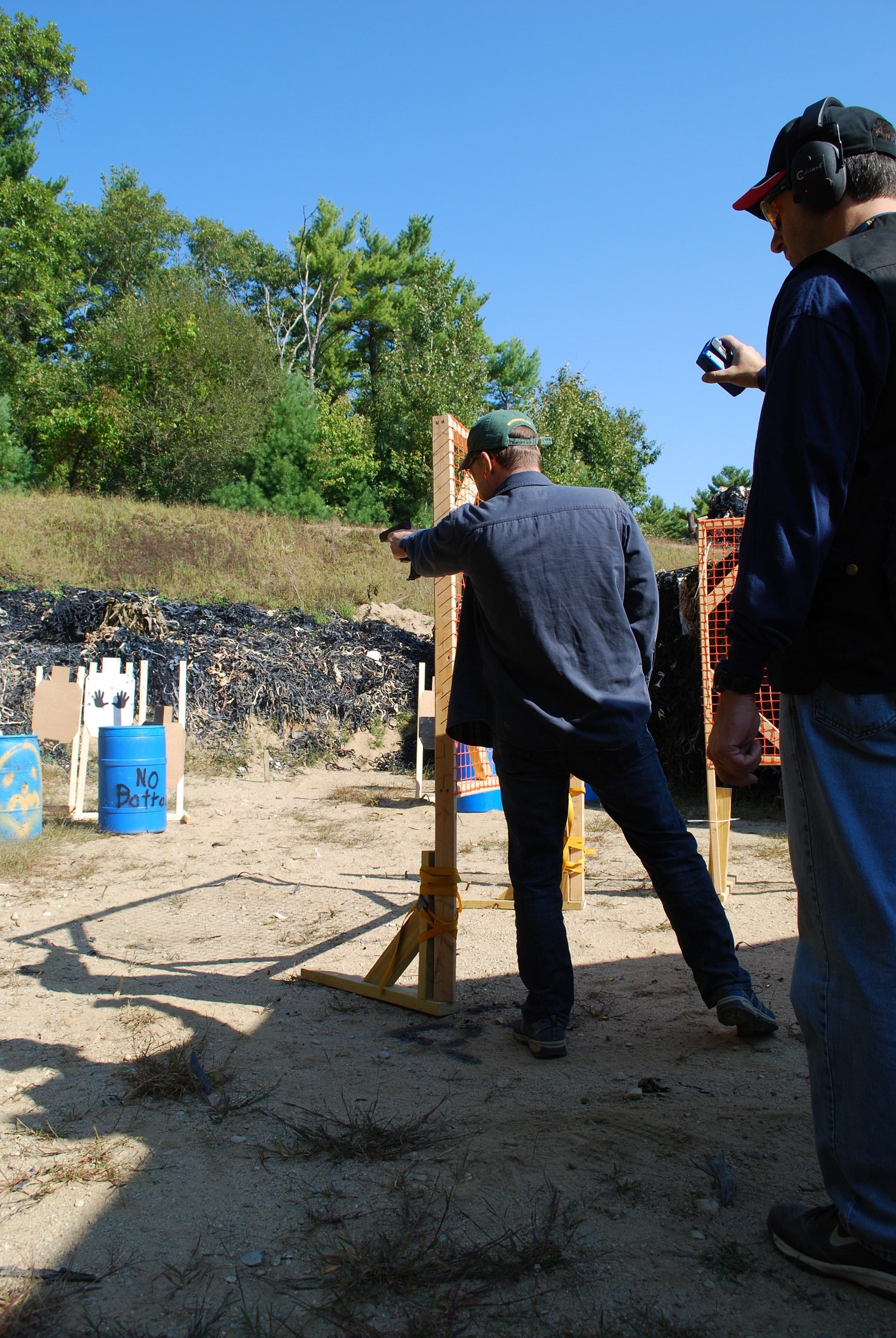 Two men at a shooting range aiming guns, one standing behind a wooden barricade and the other observing, with trees and a clear blue sky in the background.
