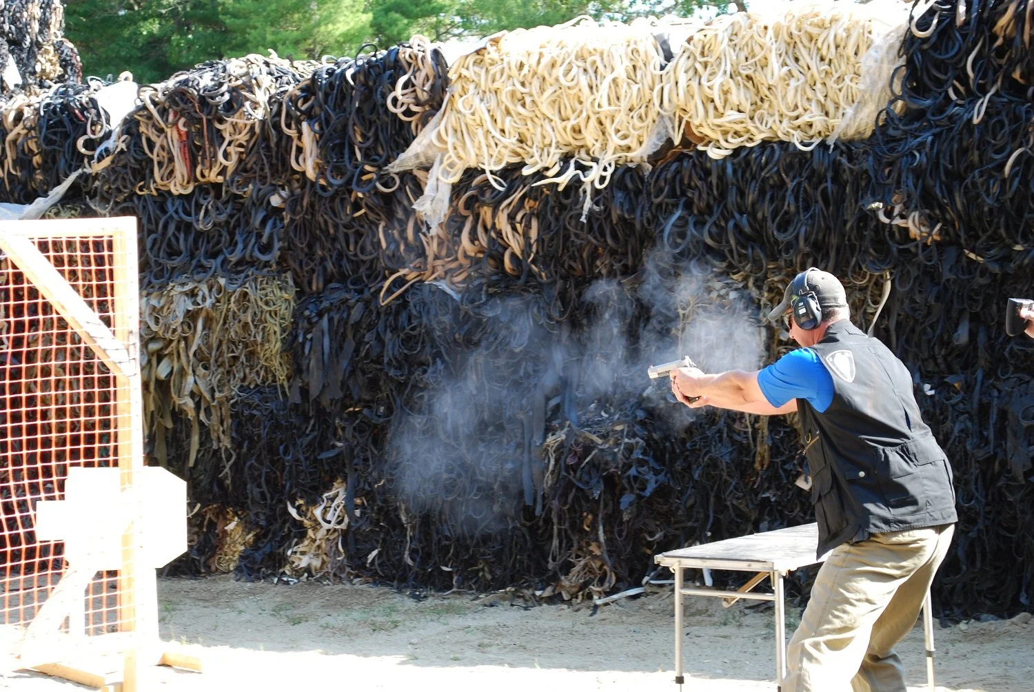 A man wearing safety ear protection and tactical gear is shooting a handgun at a shooting range, with a large pile of rubber tires in the background.