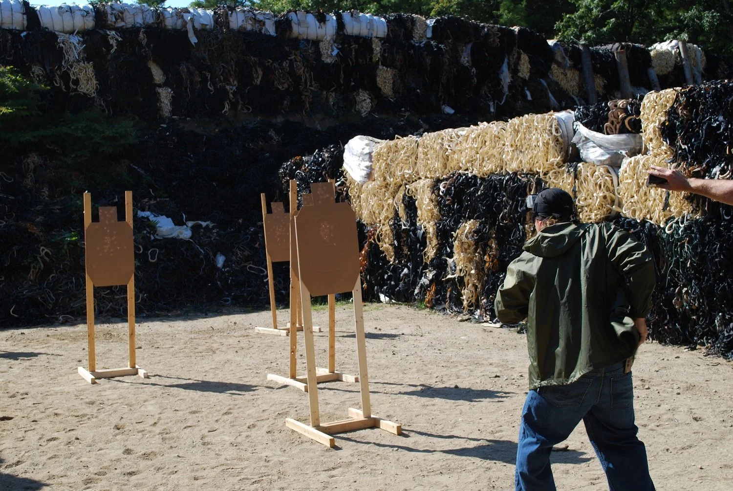 A man in a green jacket and cap practicing shooting at a training range with cardboard targets. Behind him, a large pile of ropes and plastic debris is piled against a wooded area.
