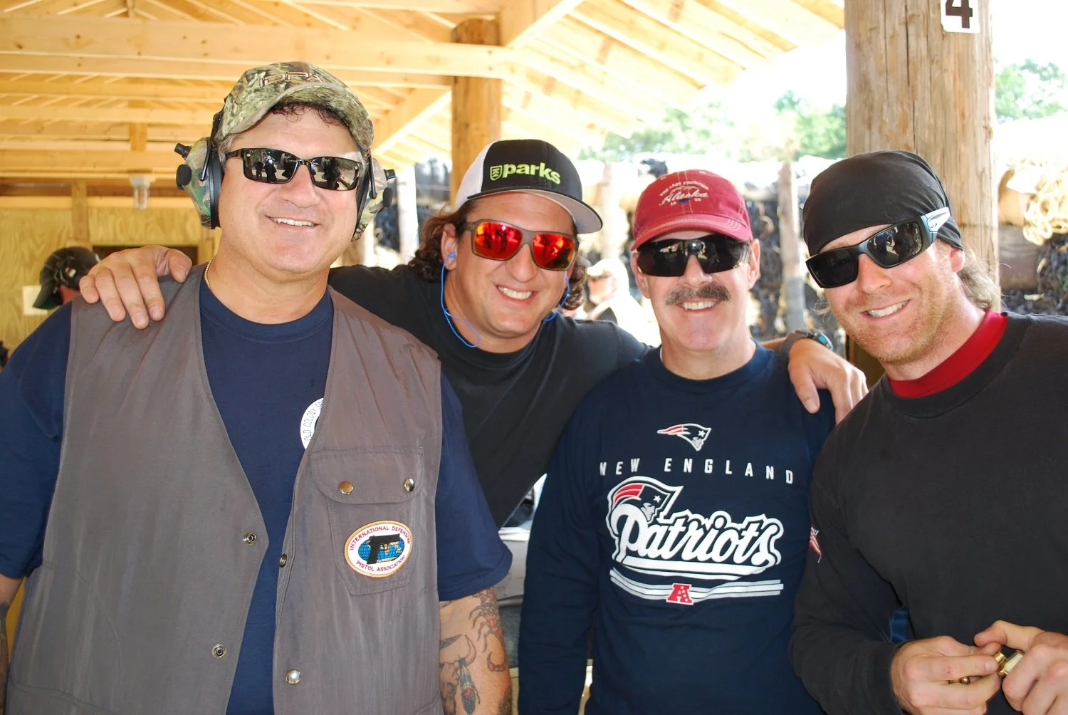 Four men smiling at a shooting range, wearing sunglasses and casual outdoor clothing, with wooden structure in the background.