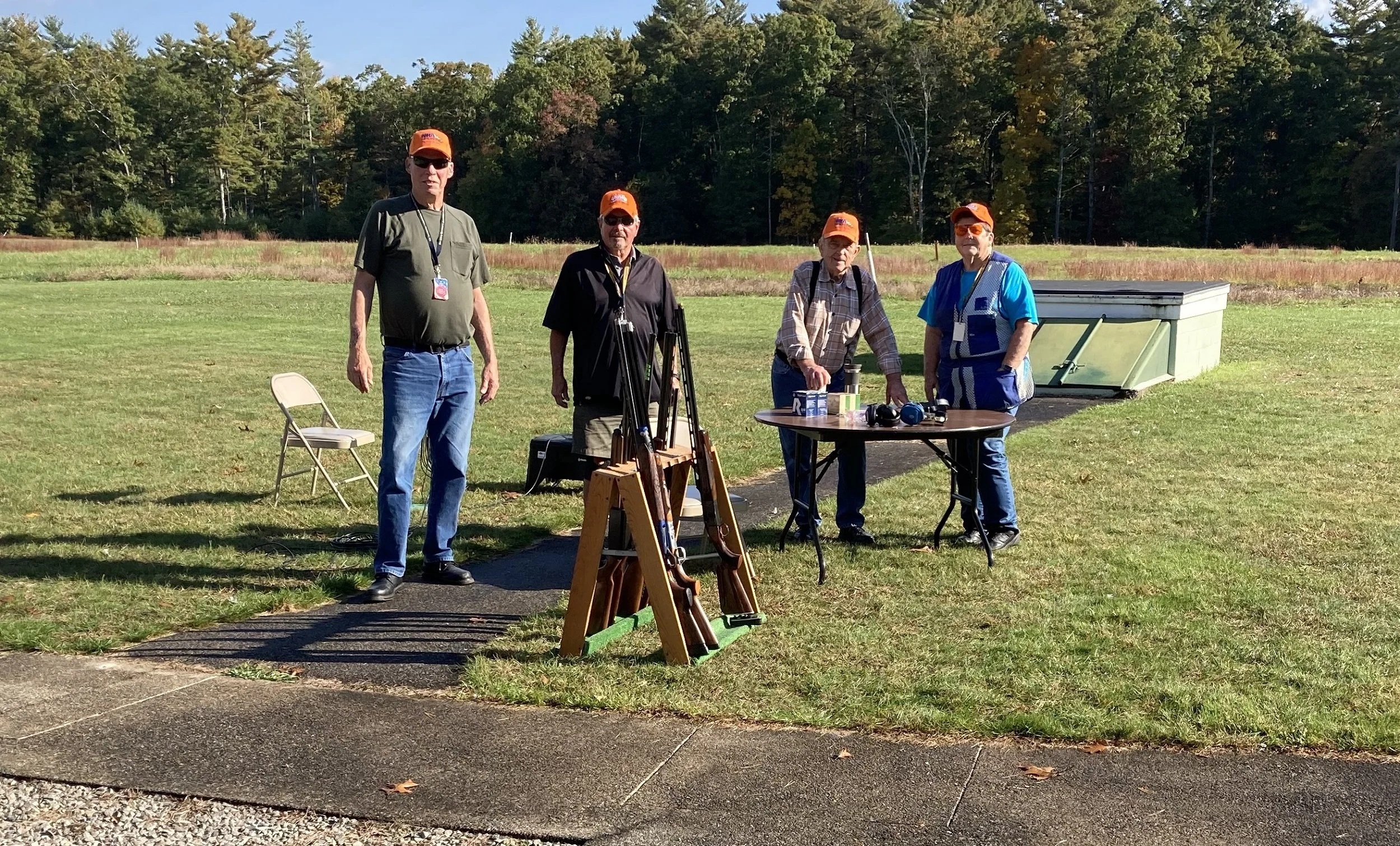 Four men standing outdoors on a grassy field with a wooded area in the background. They are wearing orange hats and standing near a folding table with items on it, and a small wooden rack holding . One man is seated, and there is a folding chair near