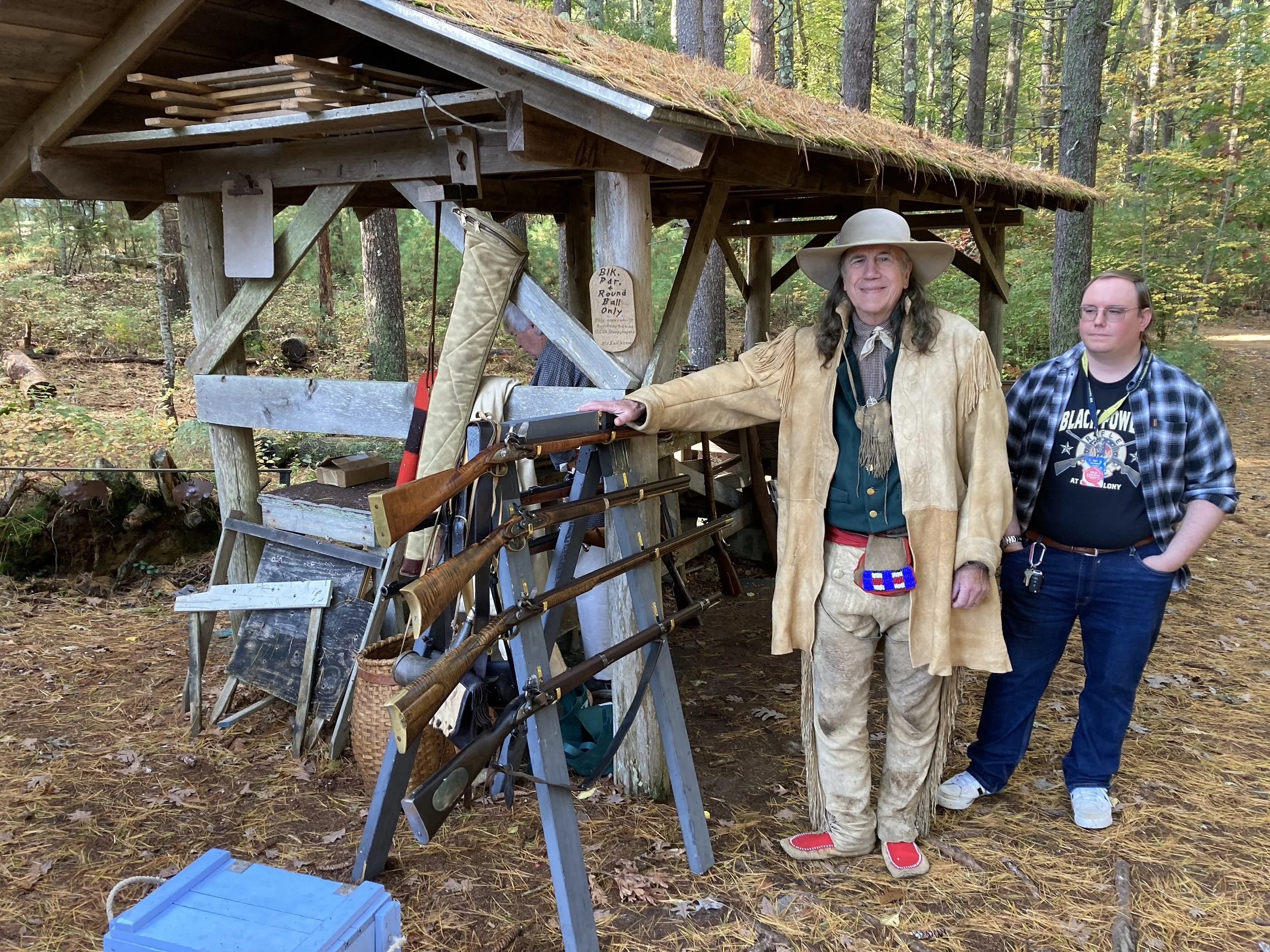 A man dressed as a historical frontiersman standing outdoors in a wooded area next to a display of vintage firearms and hunting equipment, with another person partially visible in the background.