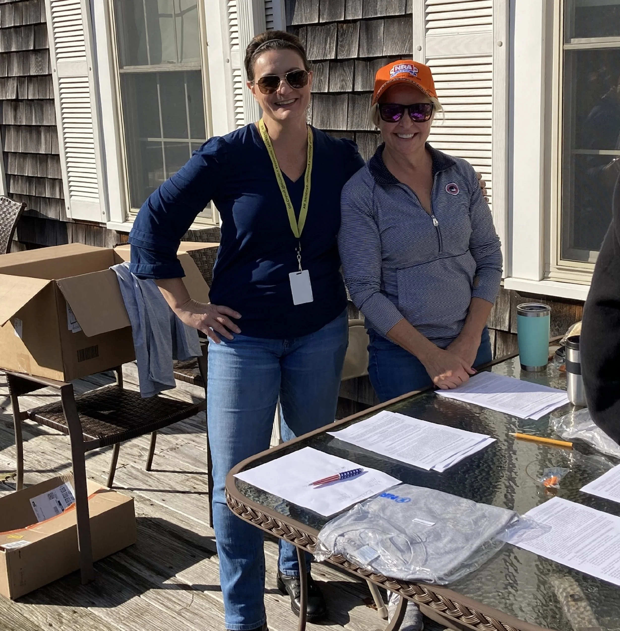 Two women standing outdoors behind a table with papers, pens, and travel mugs, smiling at the camera, with cardboard boxes nearby and a house with shingle siding in the background.