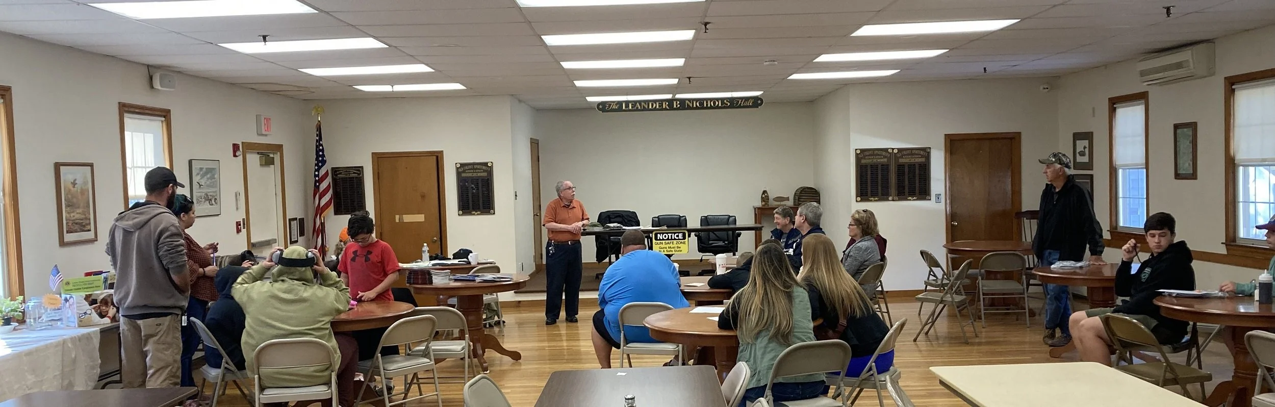 A group of people in a meeting room listening to a man speaking at the front. The room has wooden floors, framed pictures on the walls, and an American flag. Most attendees are seated at round tables, some are standing, and some are looking at their 
