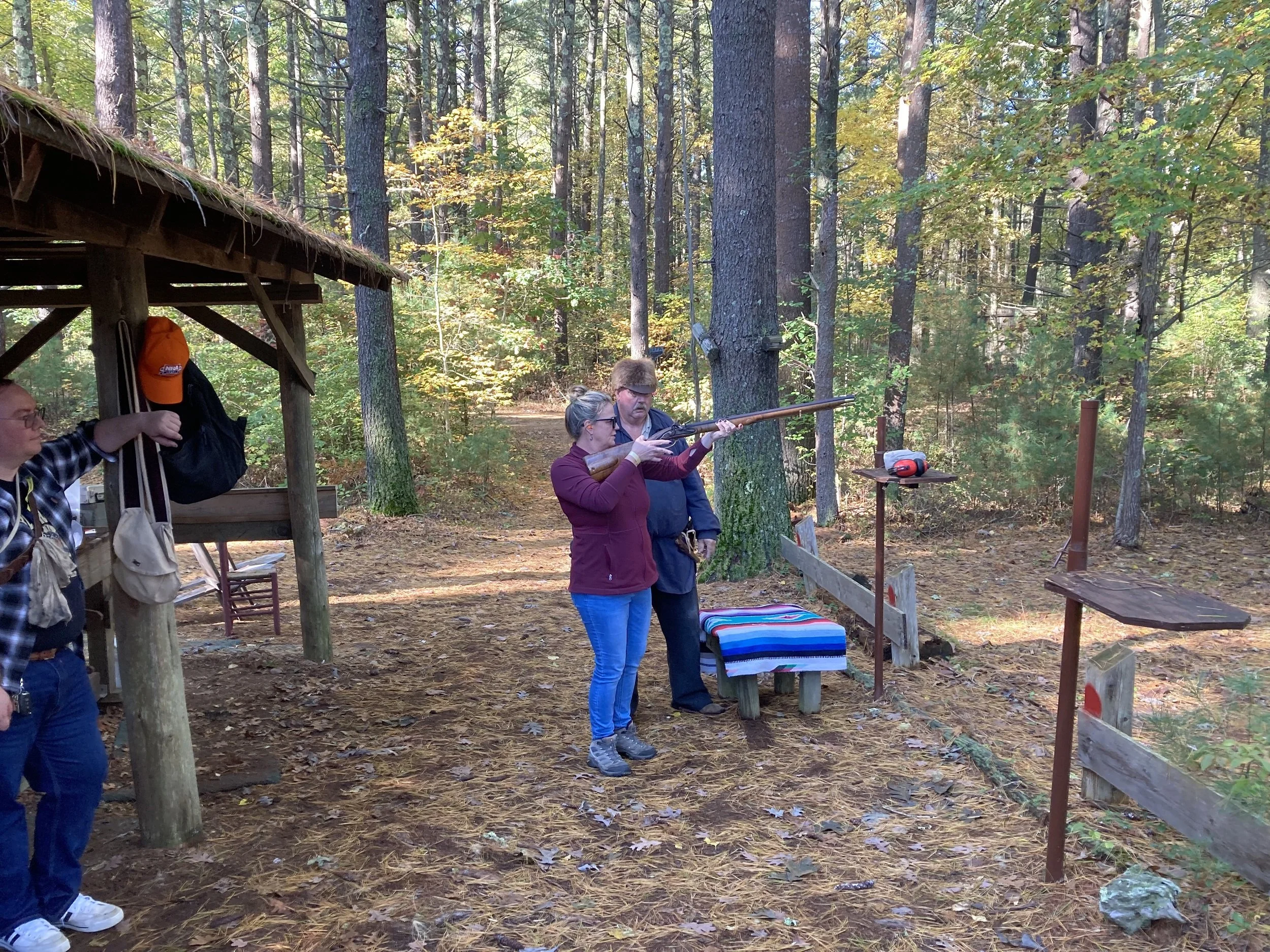 People at a shooting range in the woods, with a woman aiming a rifle under the supervision of a man, and another person standing nearby.