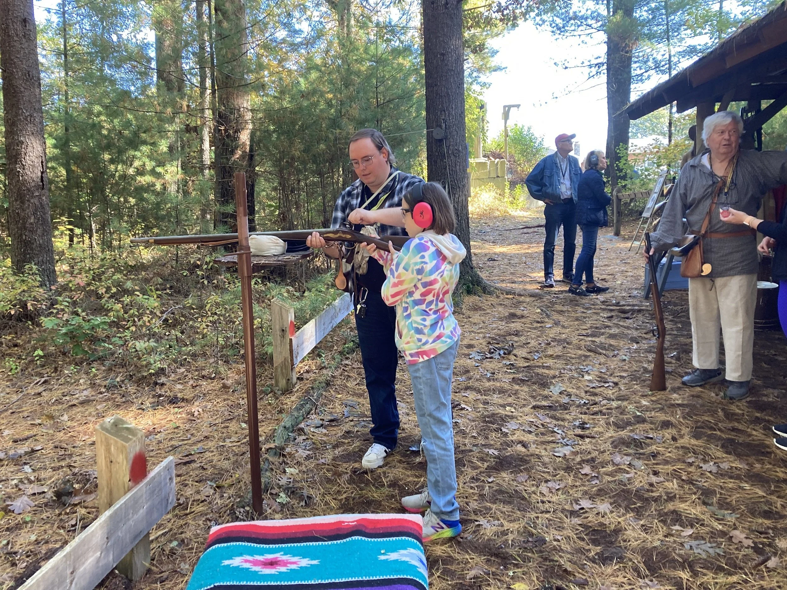 A man showing a young girl how to shoot a rifle at an outdoor shooting range surrounded by trees. Other people are in the background, some observing and some talking.