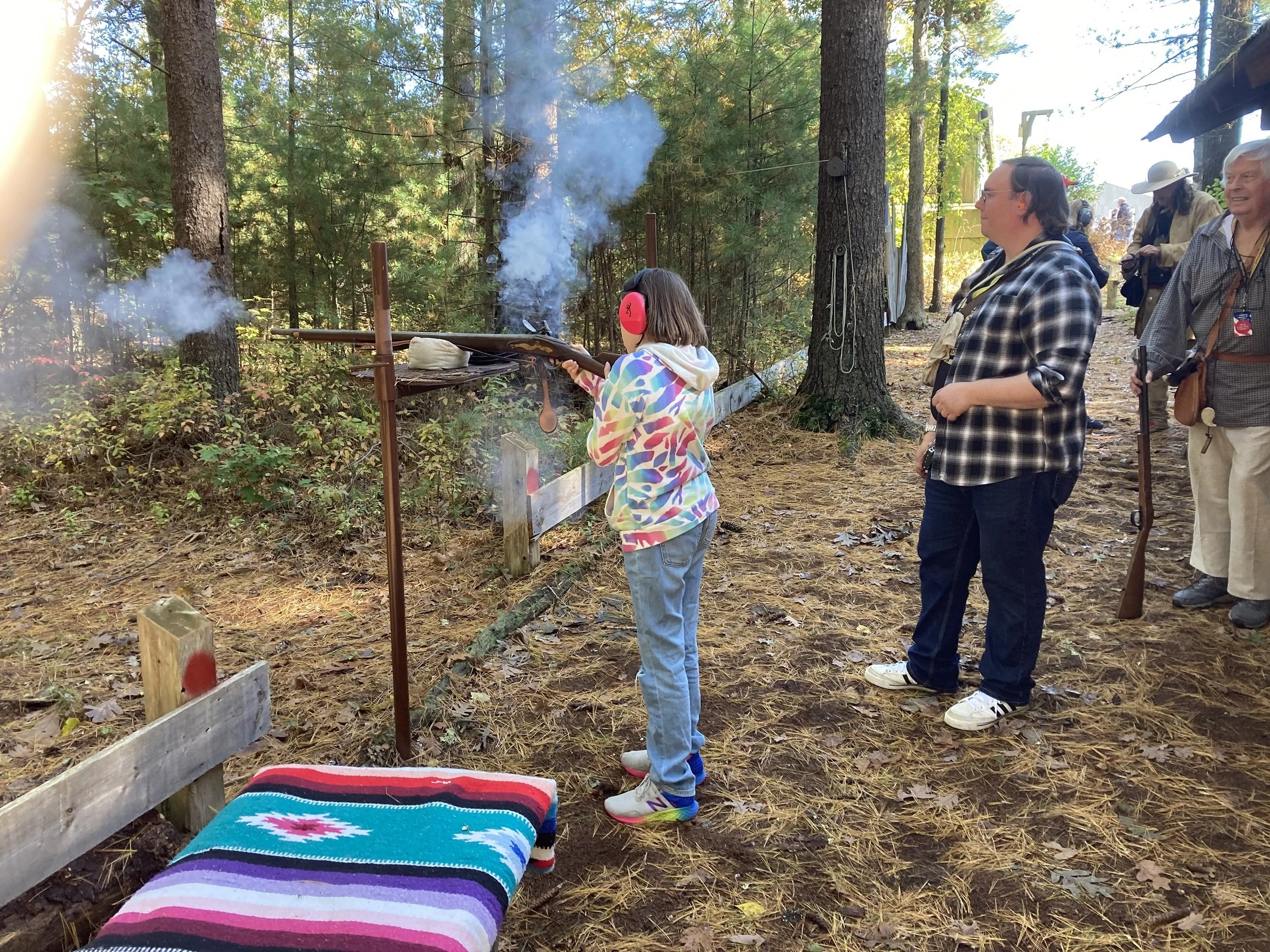A girl with headphones shooting a rifle at a shooting station in a wooded outdoor area, with other people nearby watching.