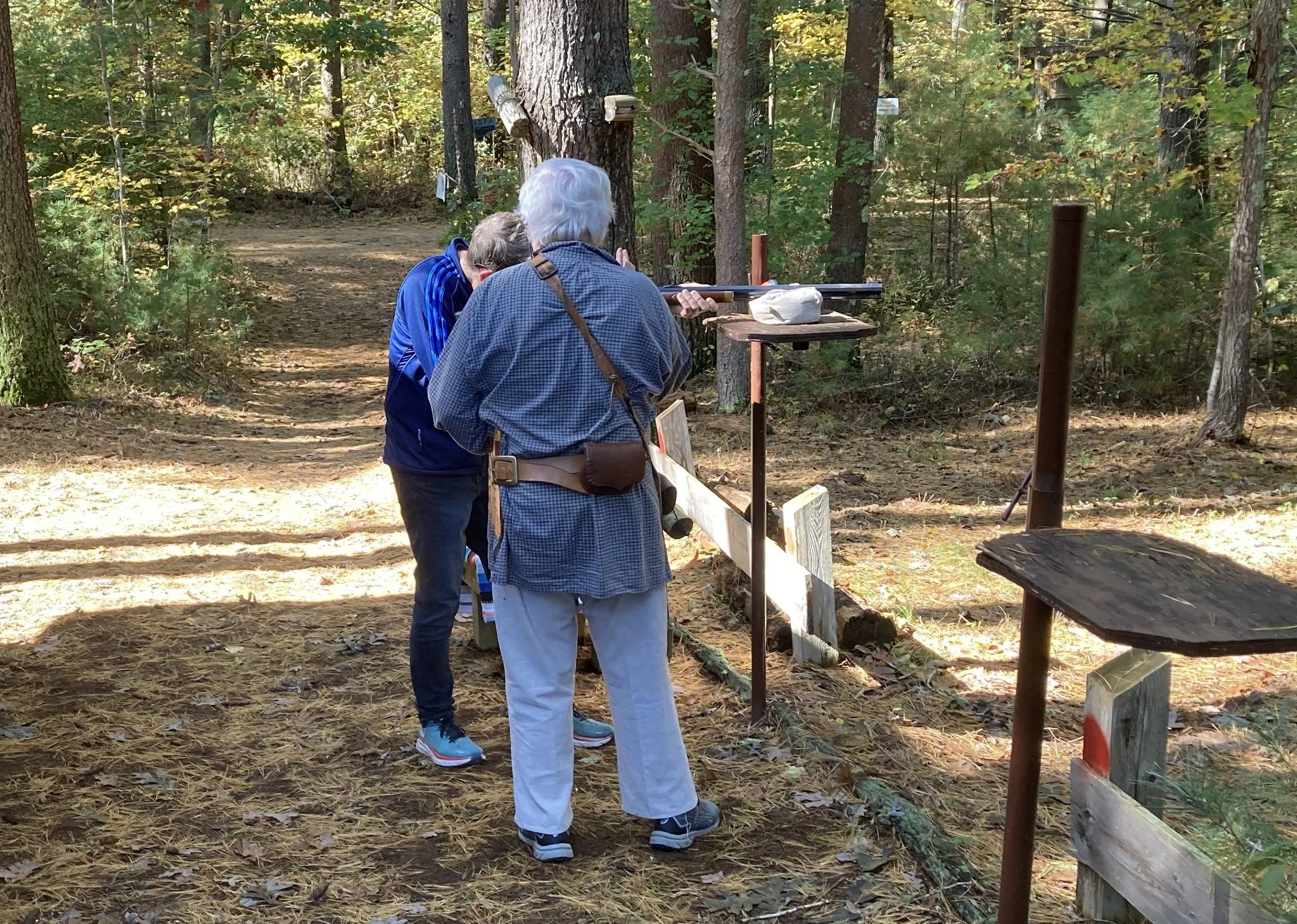 Two elderly people with white hair and gray hair shooting a rifle at a shooting range in a wooded area.