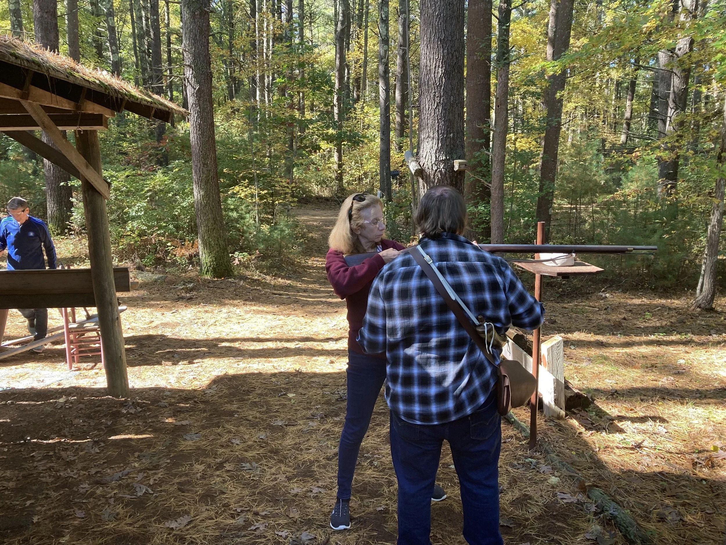 Two women are working together outdoors in a wooded area, using a rifle at a shooting station. A man is visible in the background near a pavilion.