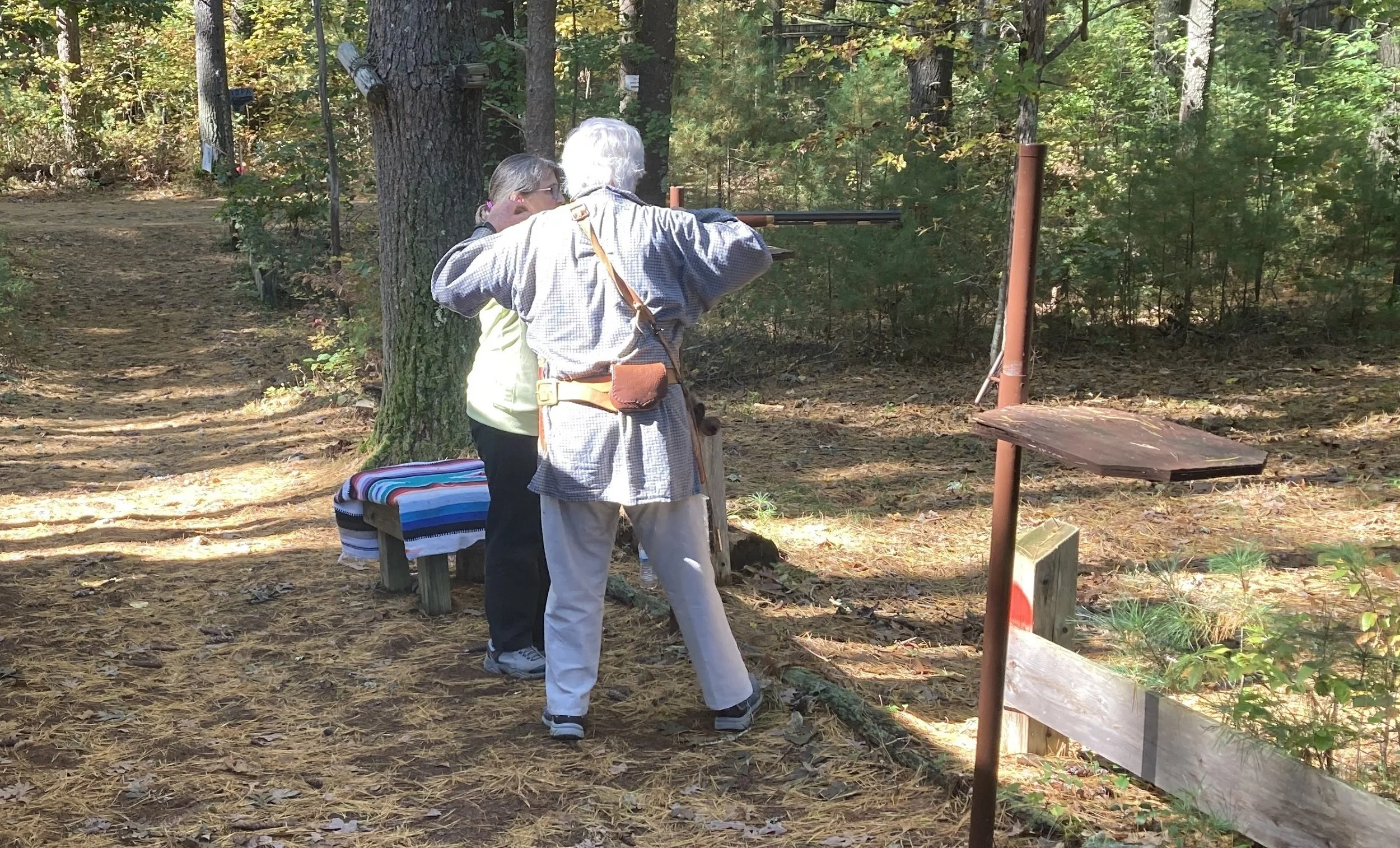 Two people, an older man and a woman, firing a rifle in a wooded outdoor shooting range.