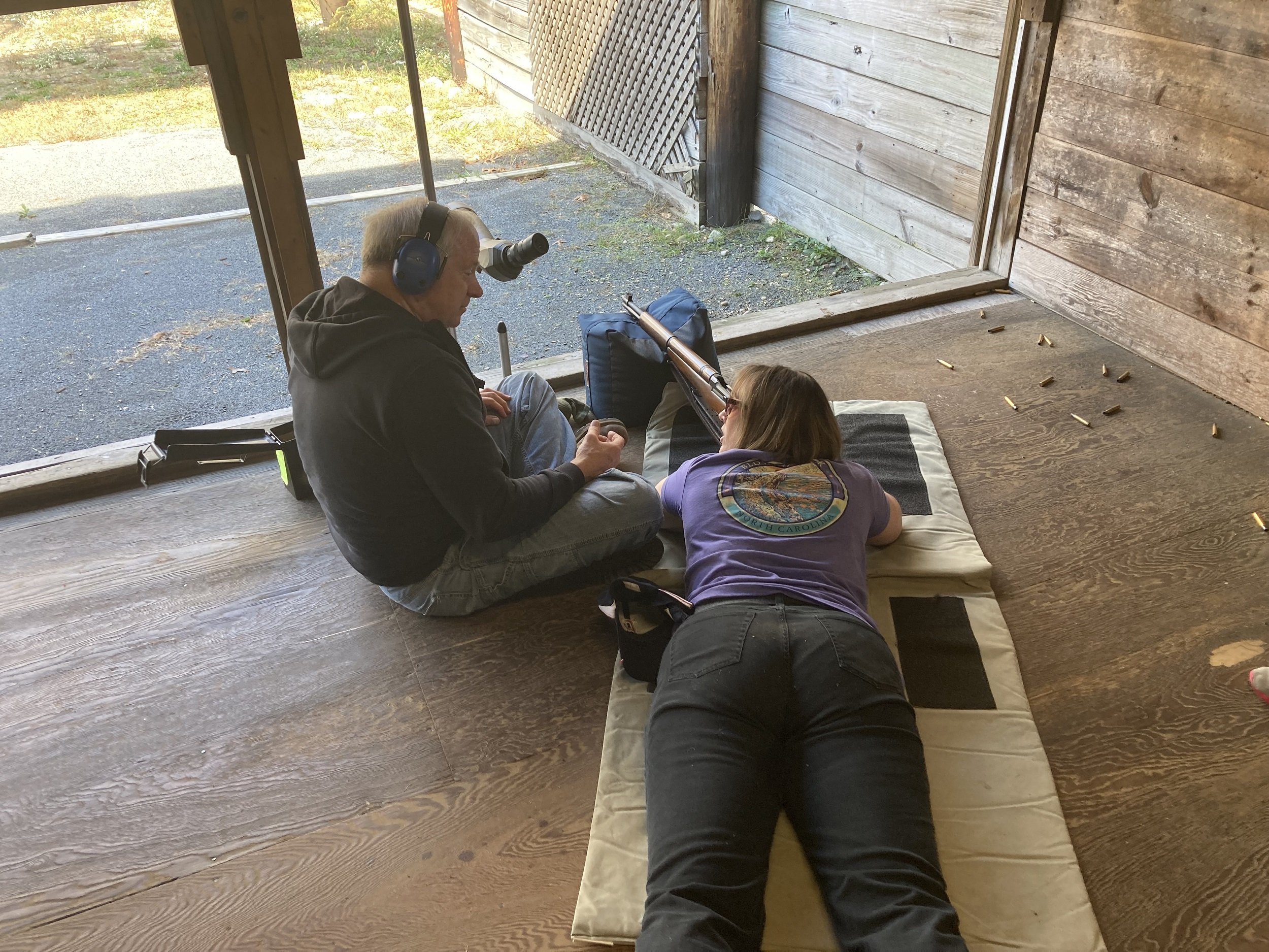 A woman lying on a pad aiming a rifle with the assistance of a man sitting next to her, in an indoor shooting range with wooden walls and no roof.