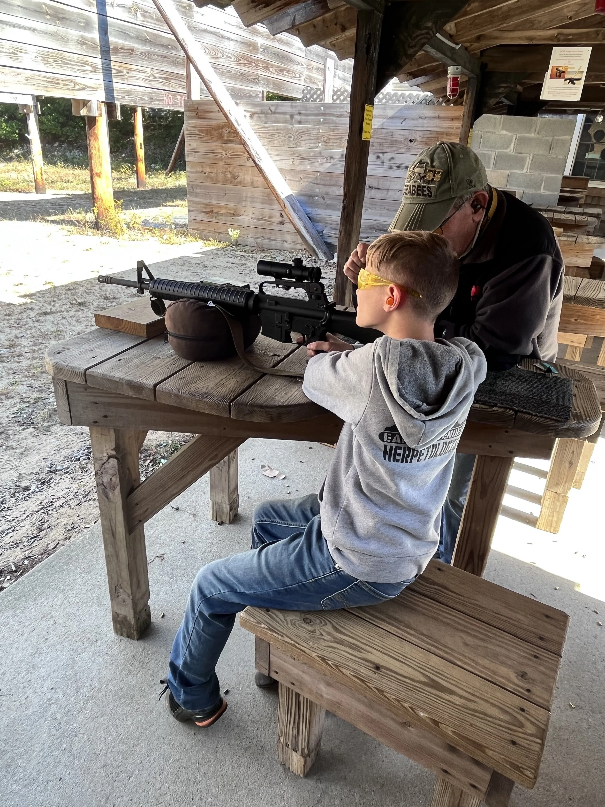 A boy with yellow safety glasses aiming a rifle supervised by an older man at an outdoor shooting range with wooden structures.