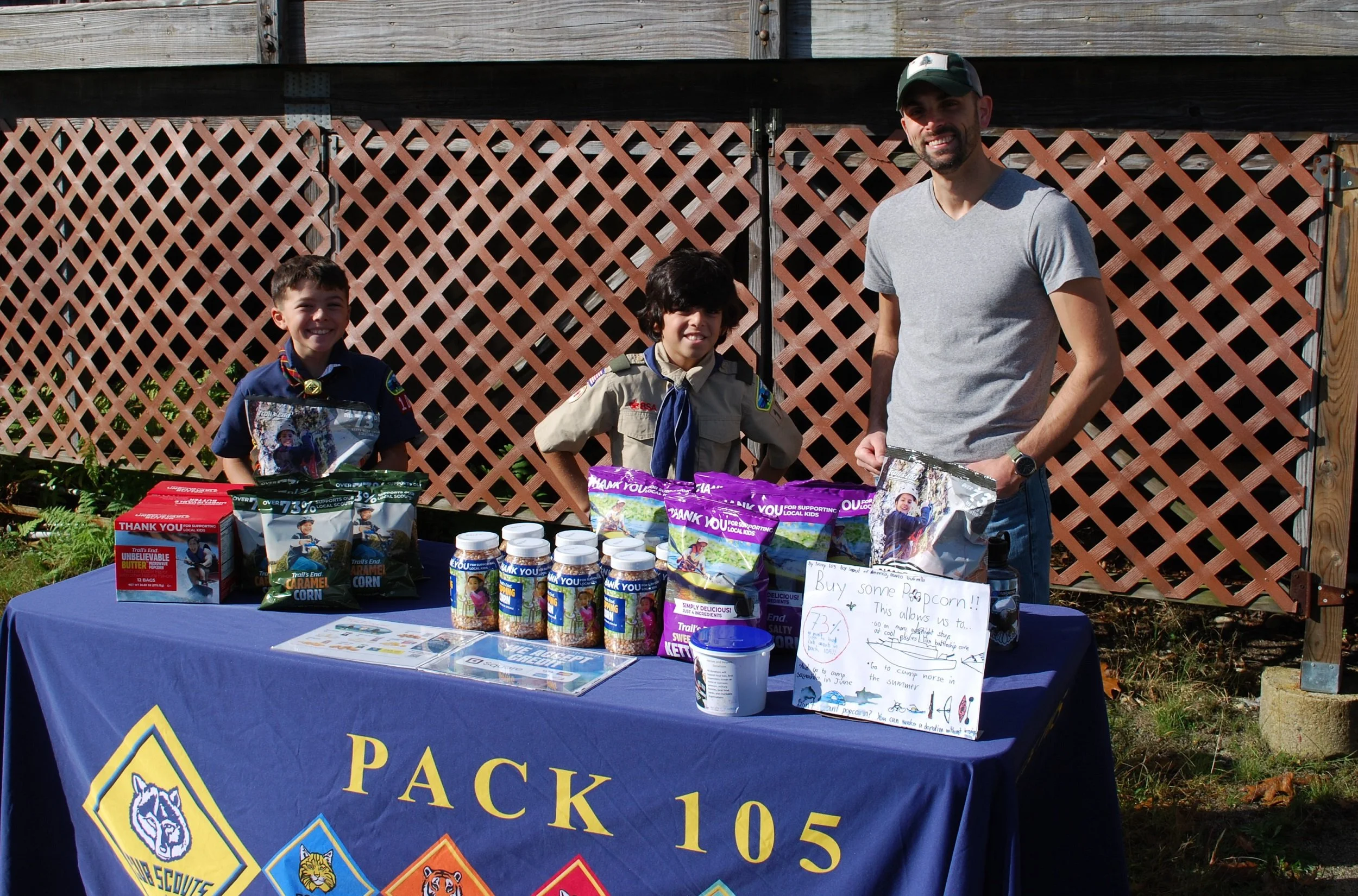 Three people standing behind a table of various snacks and drinks outdoors, with a wooden fence in the background. The tablecloth features the Boy Scouts of America logo and the text 'Pack 105'.