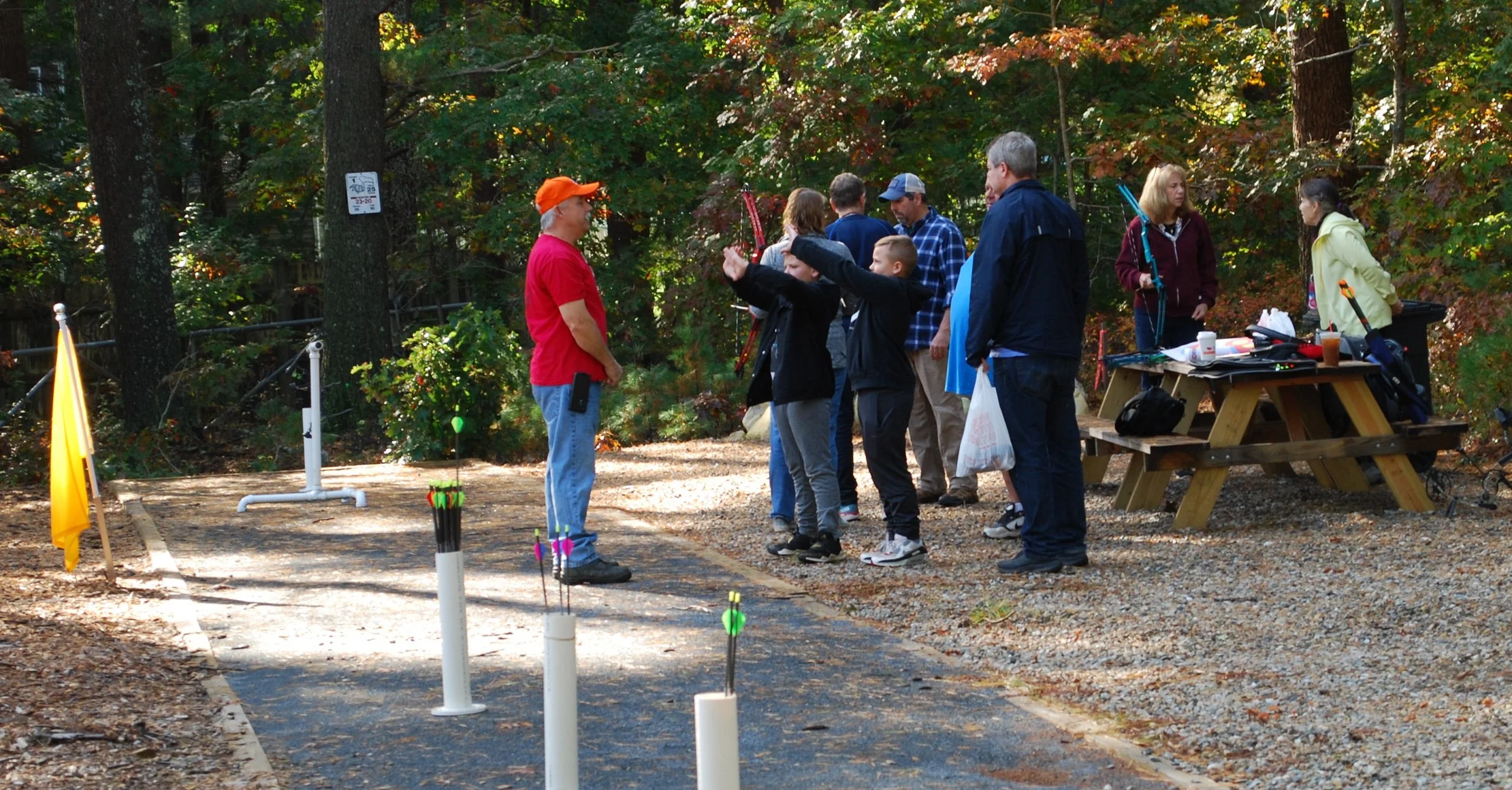 Group of people at an outdoor archery range, with children aiming bows and arrows at targets, surrounded by trees with autumn foliage.