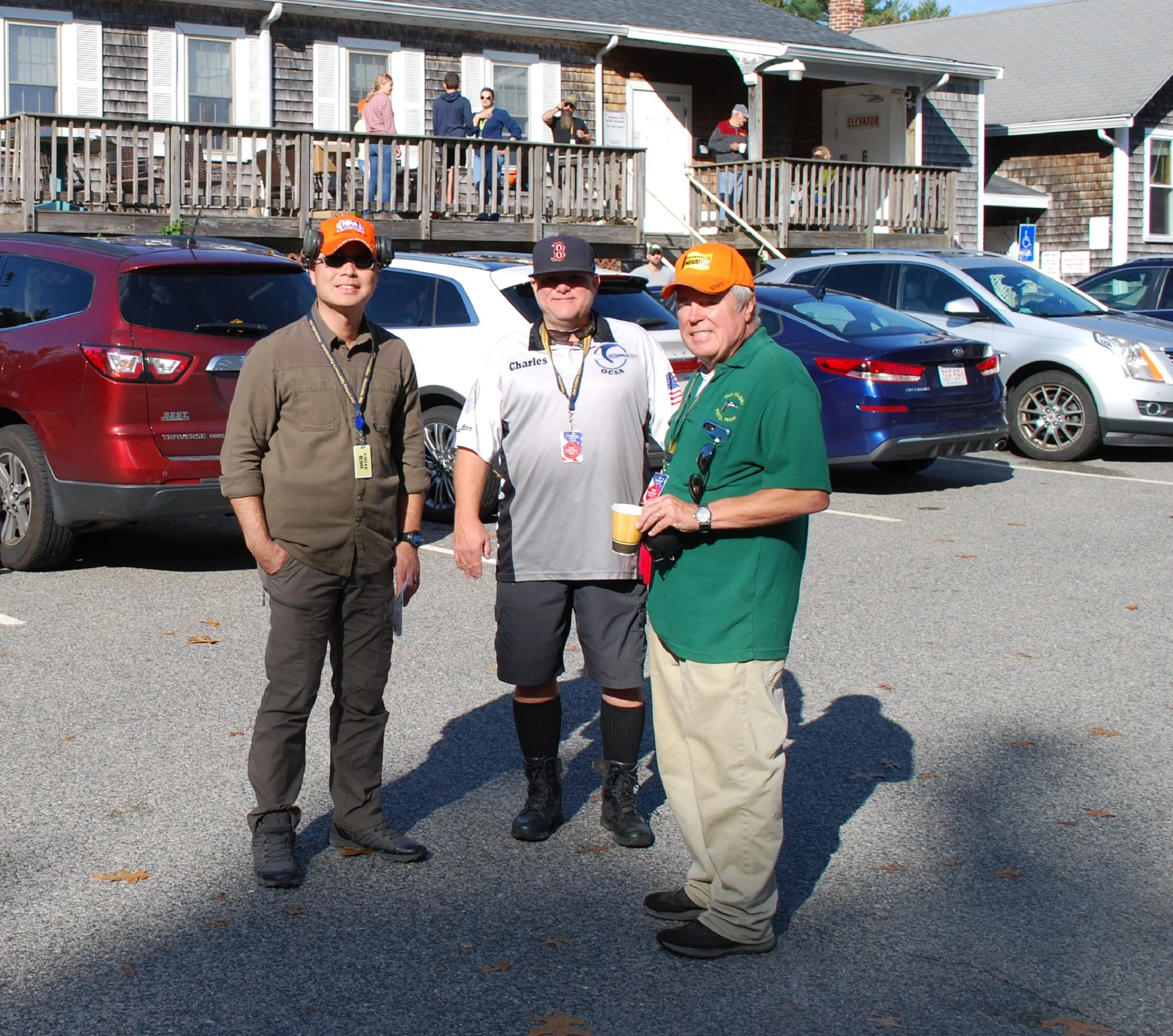 Three men standing in a parking lot, smiling, with cars and a building with a raised porch and people on it in the background.