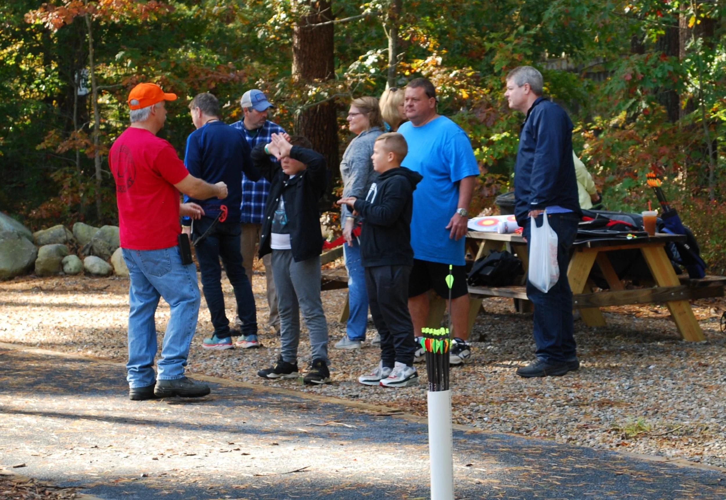 Group of people at an outdoor archery activity in a wooded area, some adjusting bows and arrows, with a man in a red shirt and orange cap instructing others.