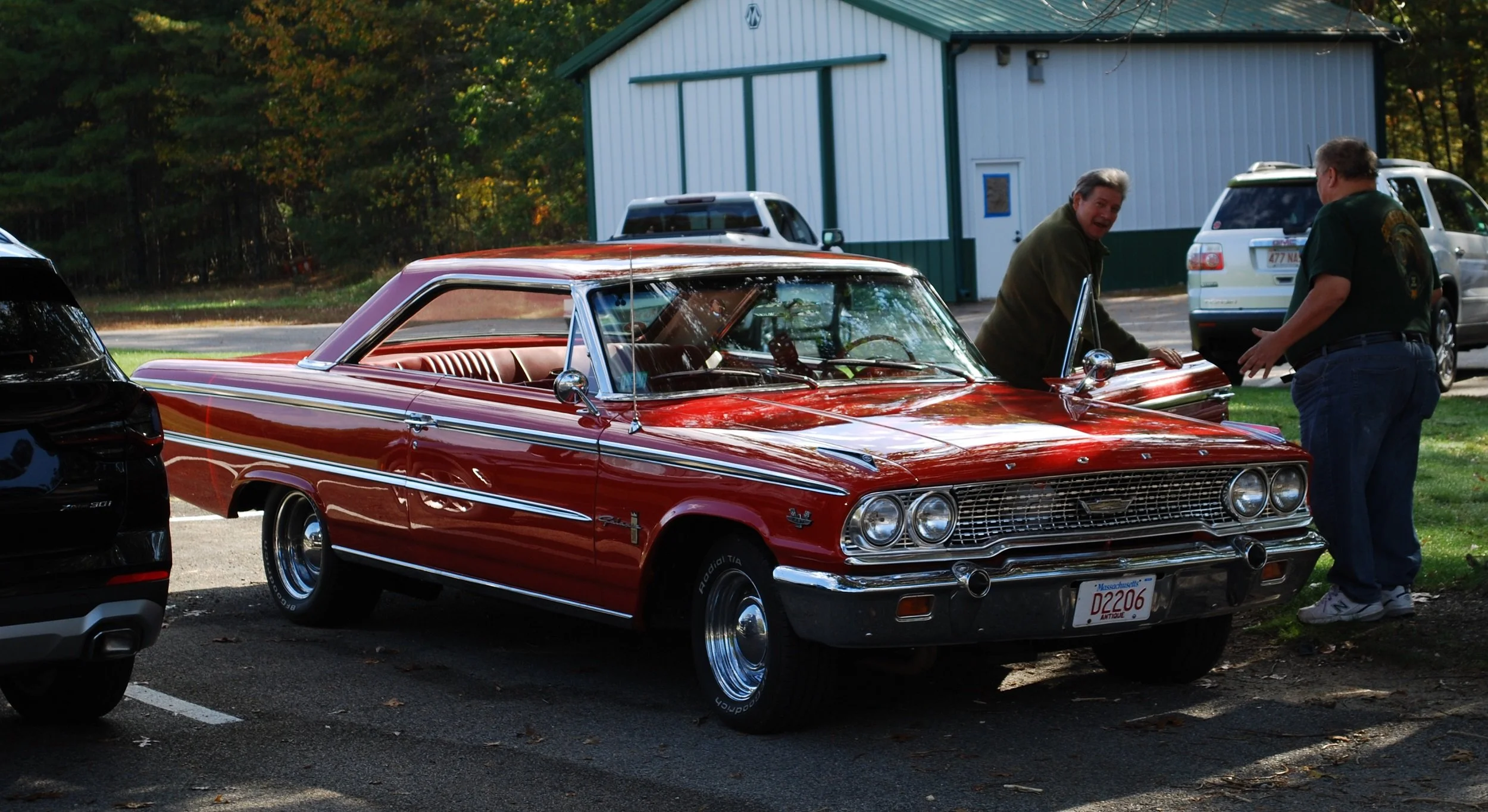 A shiny red vintage car parked in a parking lot with two men looking at it and talking, a black car on the left, and other cars and trees in the background.