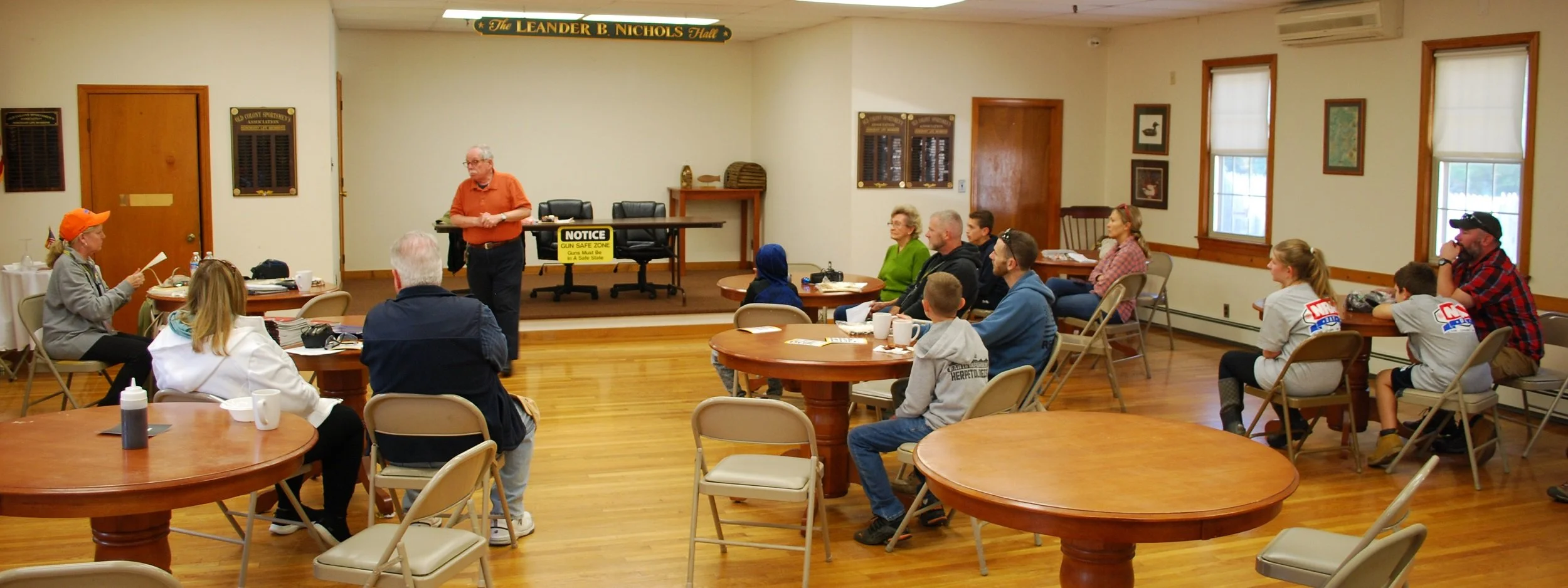 A group of people attending a meeting or workshop in a room with wooden floors and walls. There is a man standing at the front, speaking to the audience, who are seated at round tables. The room has windows with blinds, and a sign that reads 'The Lea