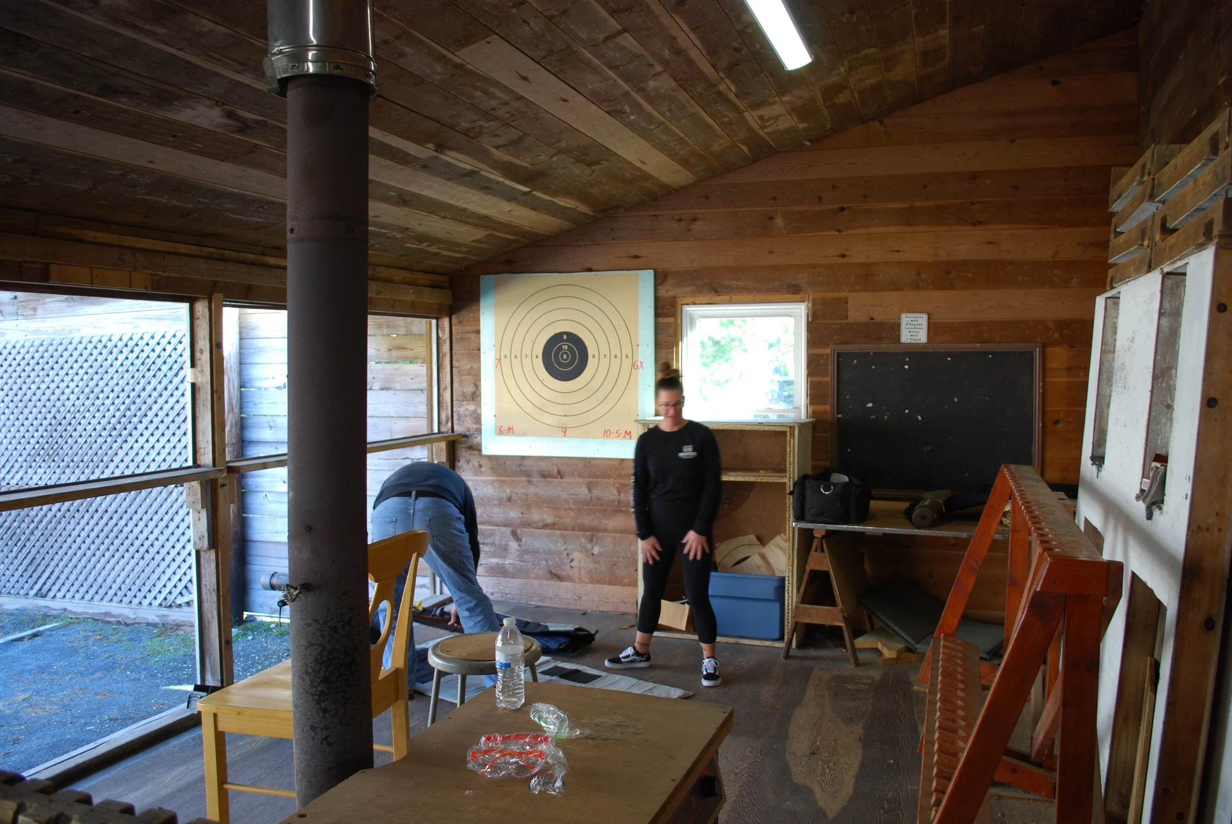 Wooden interior of a shooting range with a target on the wall, two people preparing or cleaning gear, a water bottle on the table, and various items on shelves and the floor.