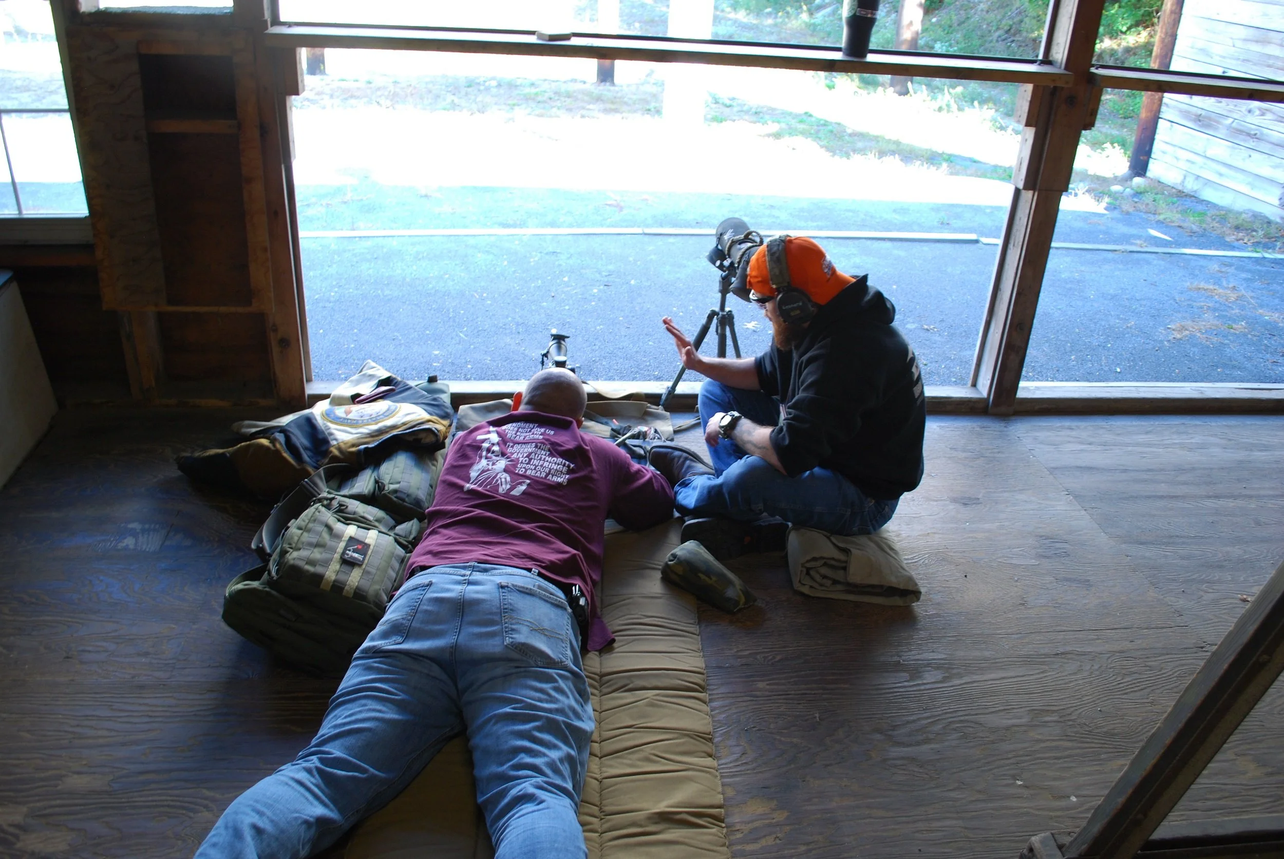 Two men are working with a spotting scope and tripod inside a wooden building, with one man sitting and the other lying face down on a cushion, near a large open wall.