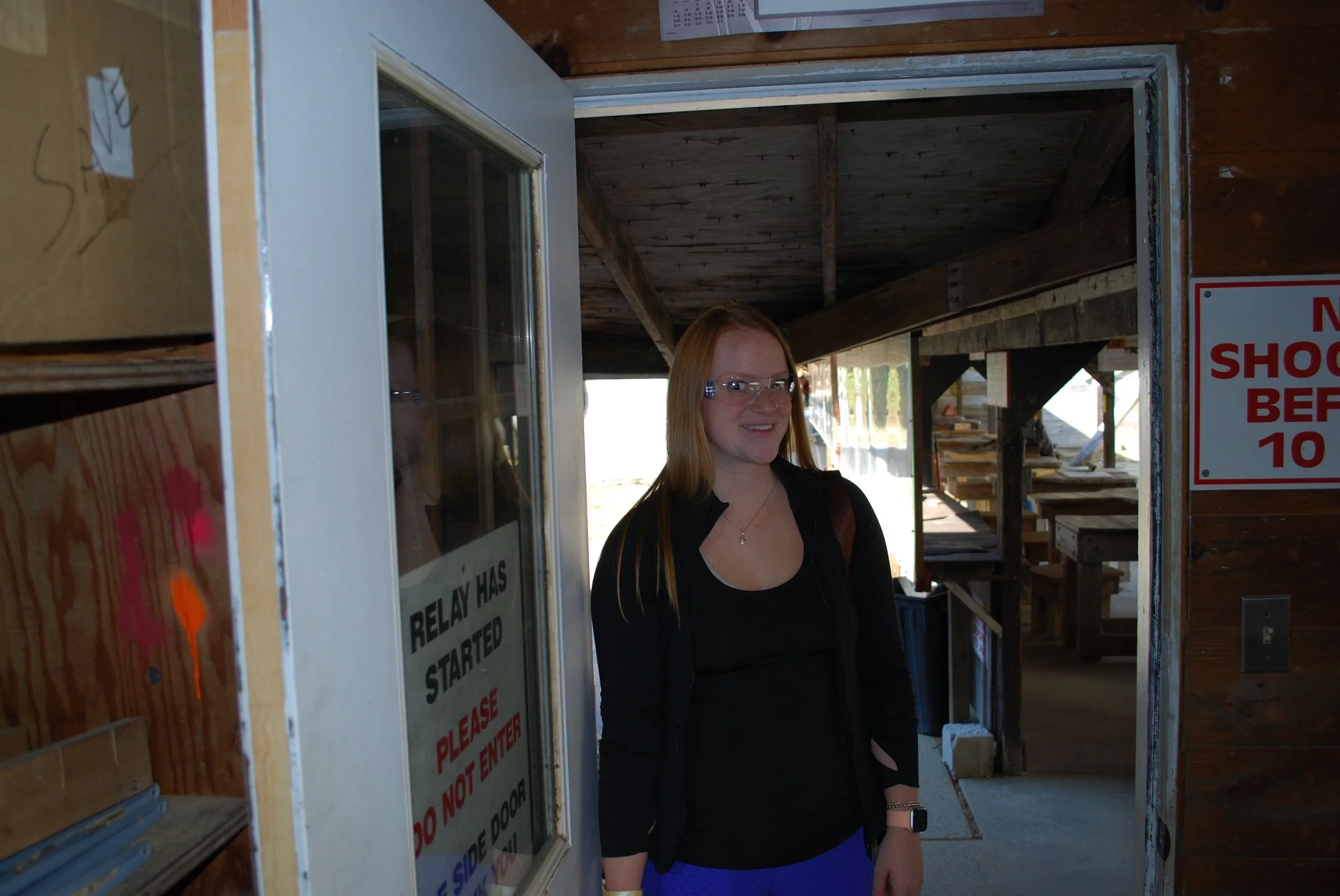 A woman with red hair wearing glasses, a black top, and a black jacket standing at a door in a wooden building, smiling.