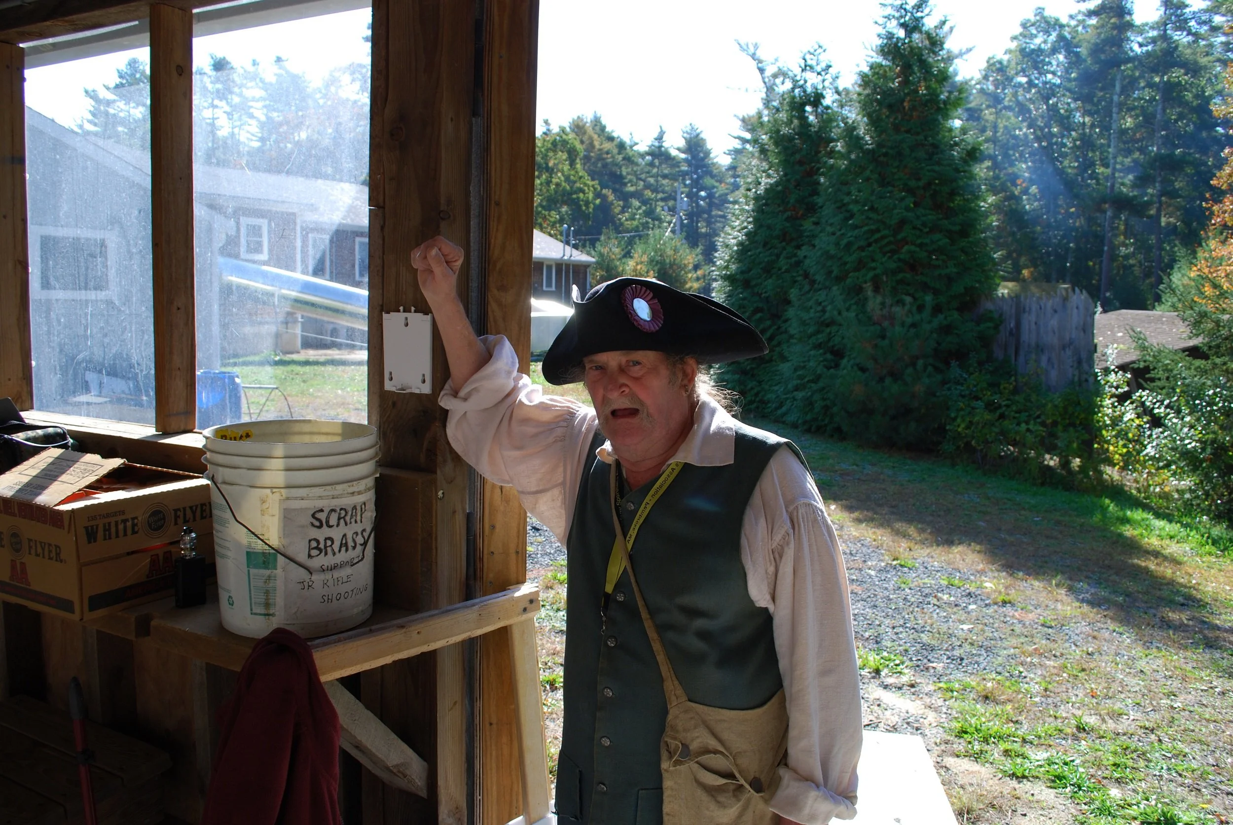 Man dressed as a pirate wearing a large pirate hat with a badge, inside a wooden shed, raising one arm, with outdoor trees and neighborhood visible through the open door