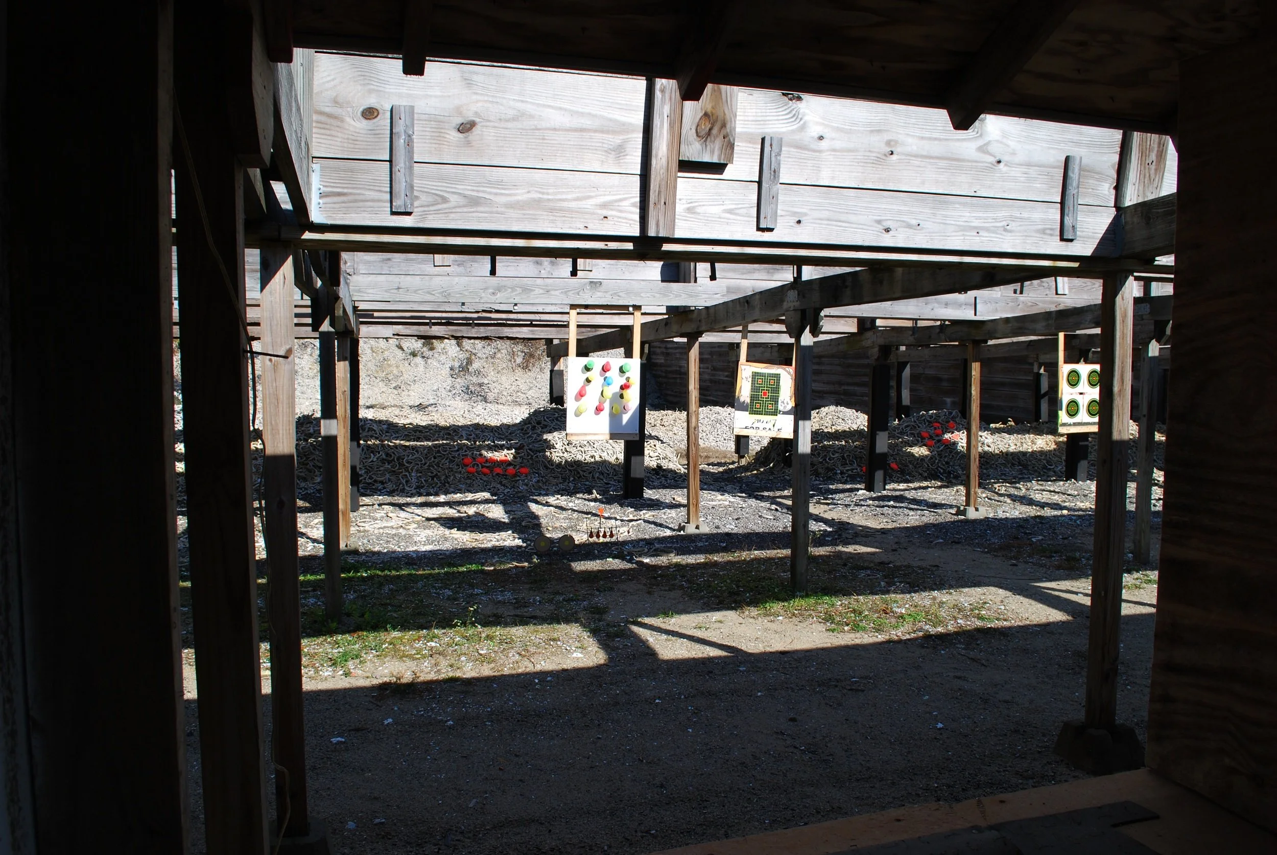 Outdoor shooting range with multiple targets set up along a gravel ground, viewed from beneath a wooden shelter.