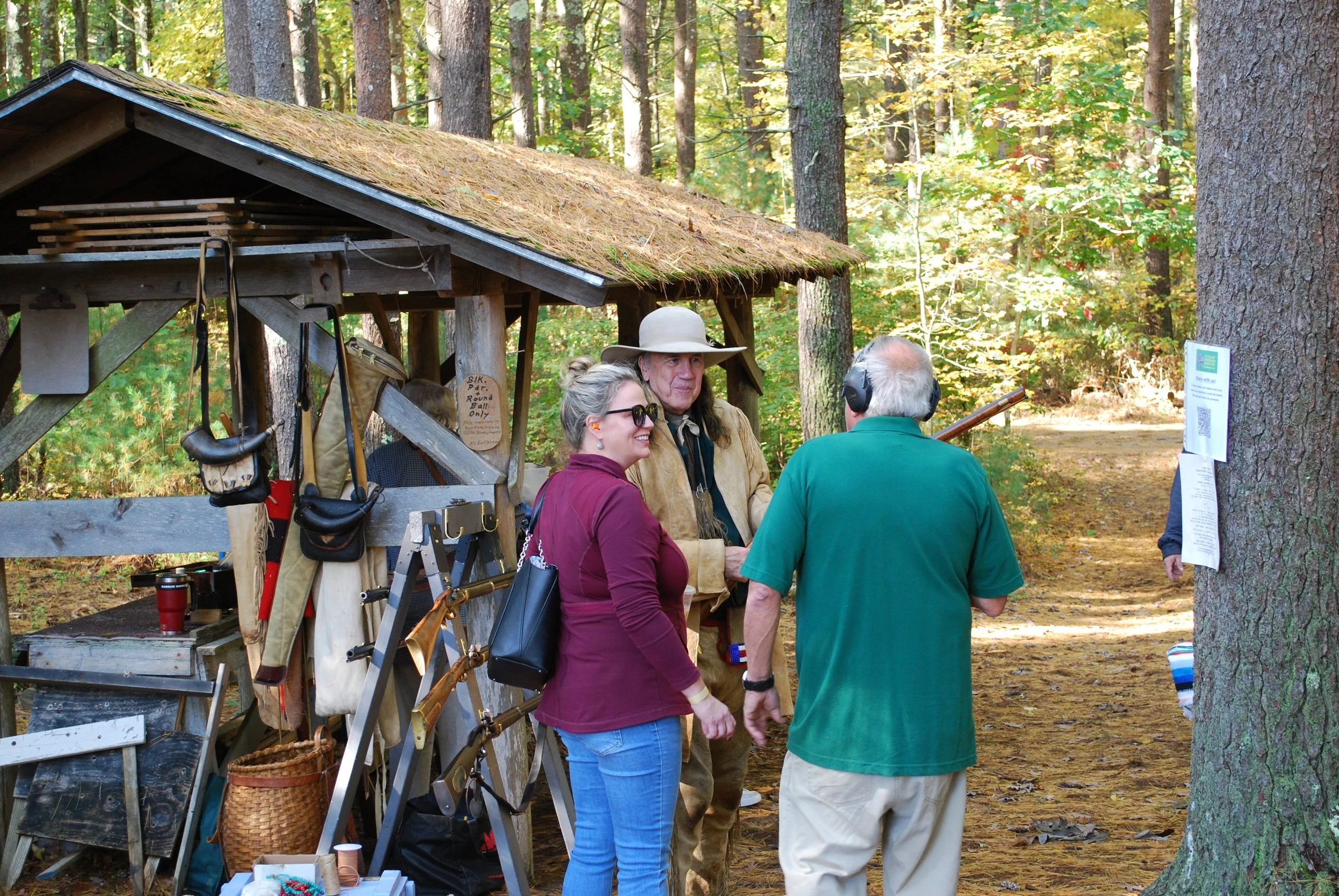 People talking at outdoor gun range with various firearms and gear displayed under wooden shelter amidst trees