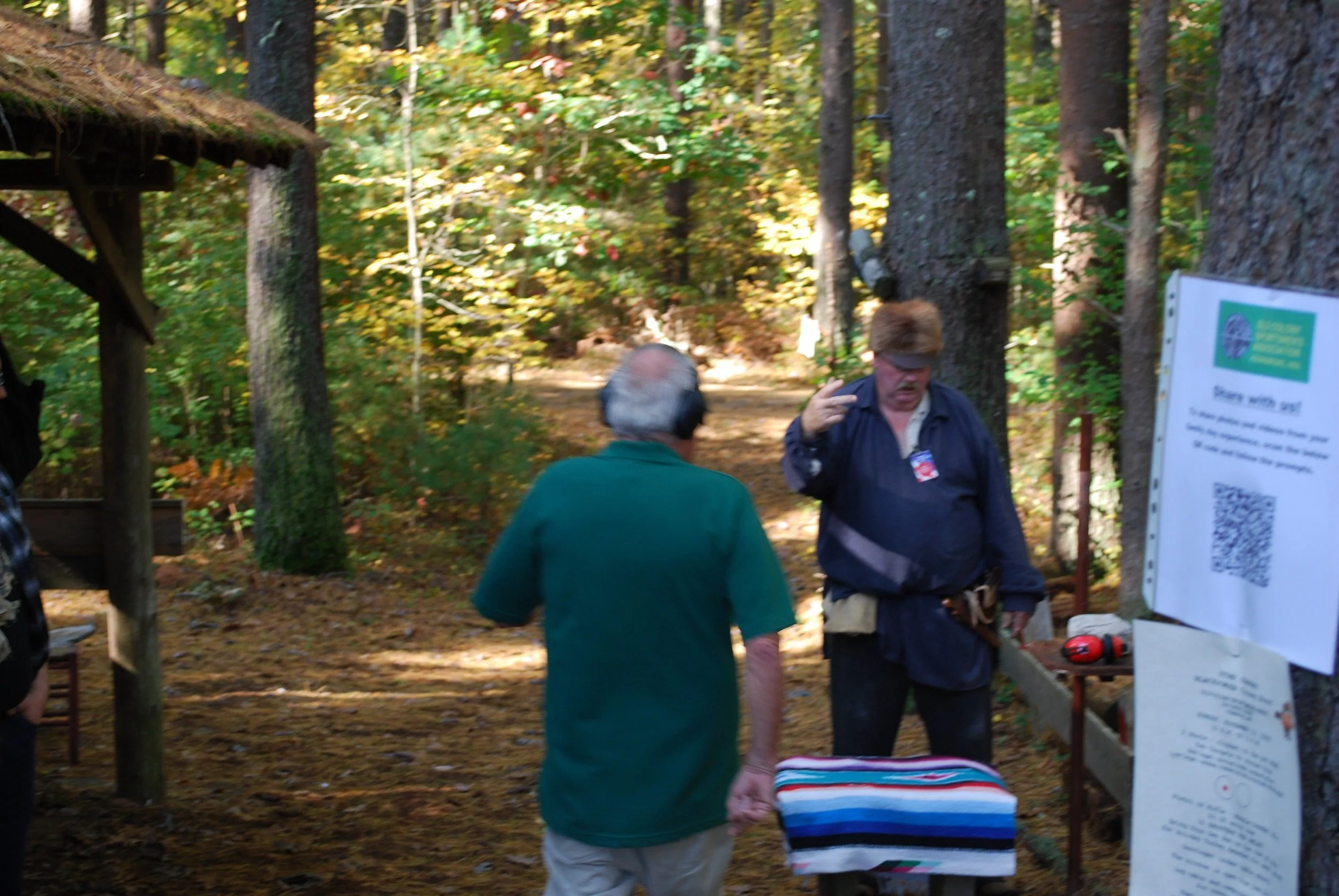Two men talking outdoors in a wooded area, with a sign and some equipment nearby.
