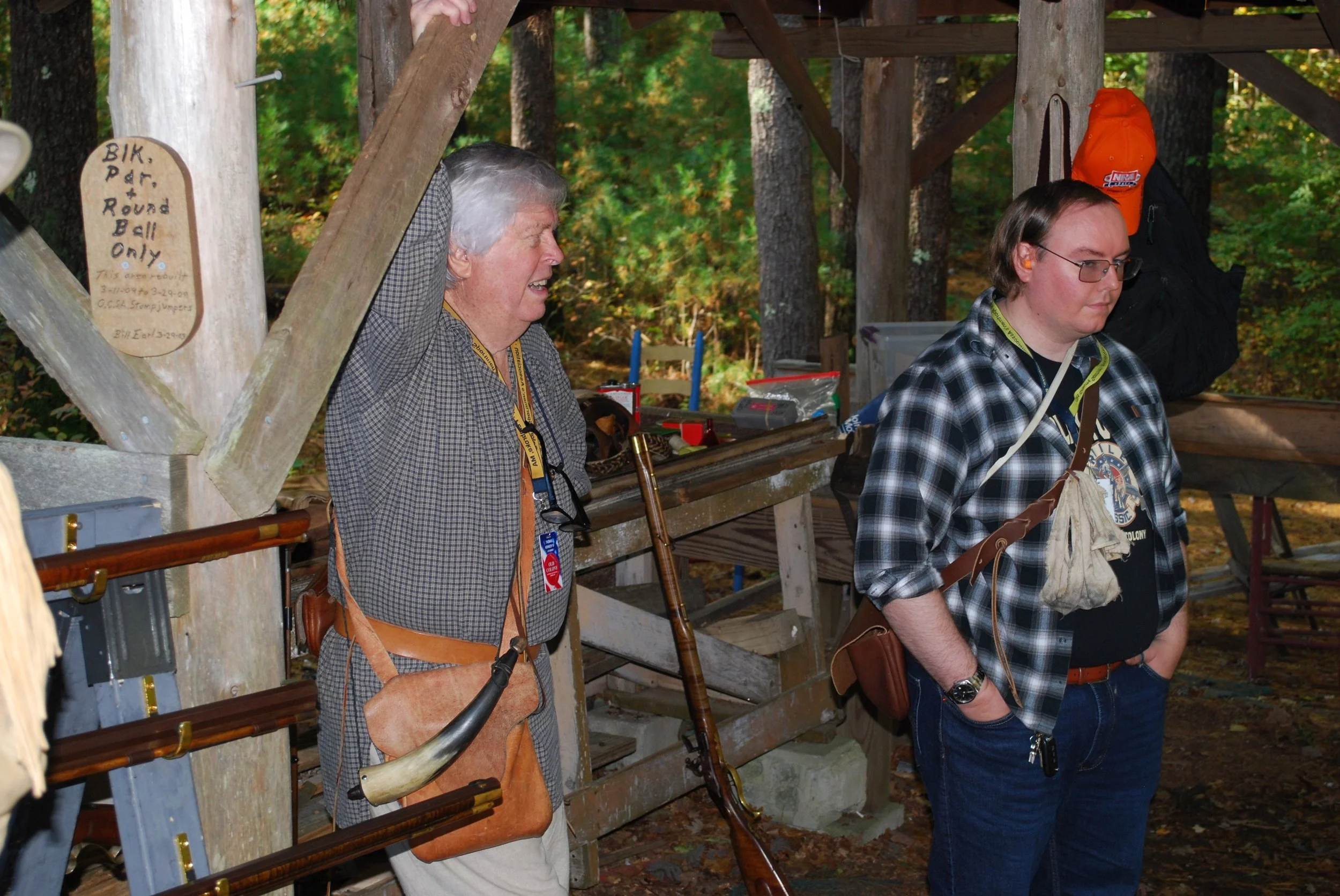 Two men standing outdoors under a wooden structure, with trees in the background. The man on the left is elderly with gray hair, wearing a checked shirt, carrying a leather pouch, and next to an antique rifle. The man on the right is younger, wearing