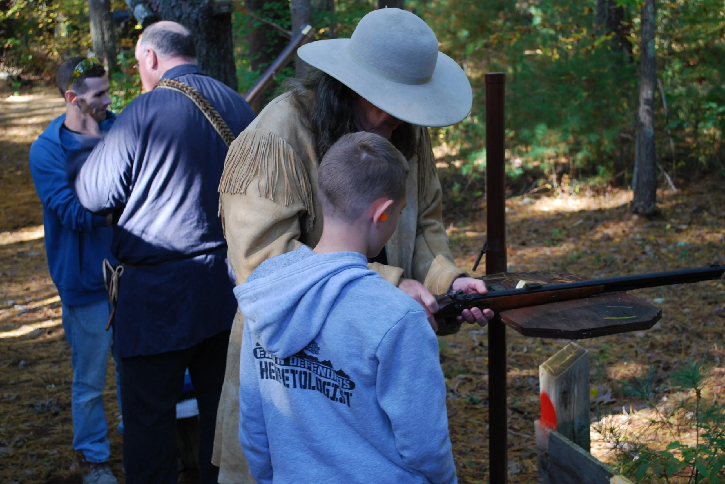 A man in a beige fringed jacket and gray wide-brimmed hat teaches a boy in a blue hoodie how to shoot a rifle outdoors in a wooded area, with two men in the background.