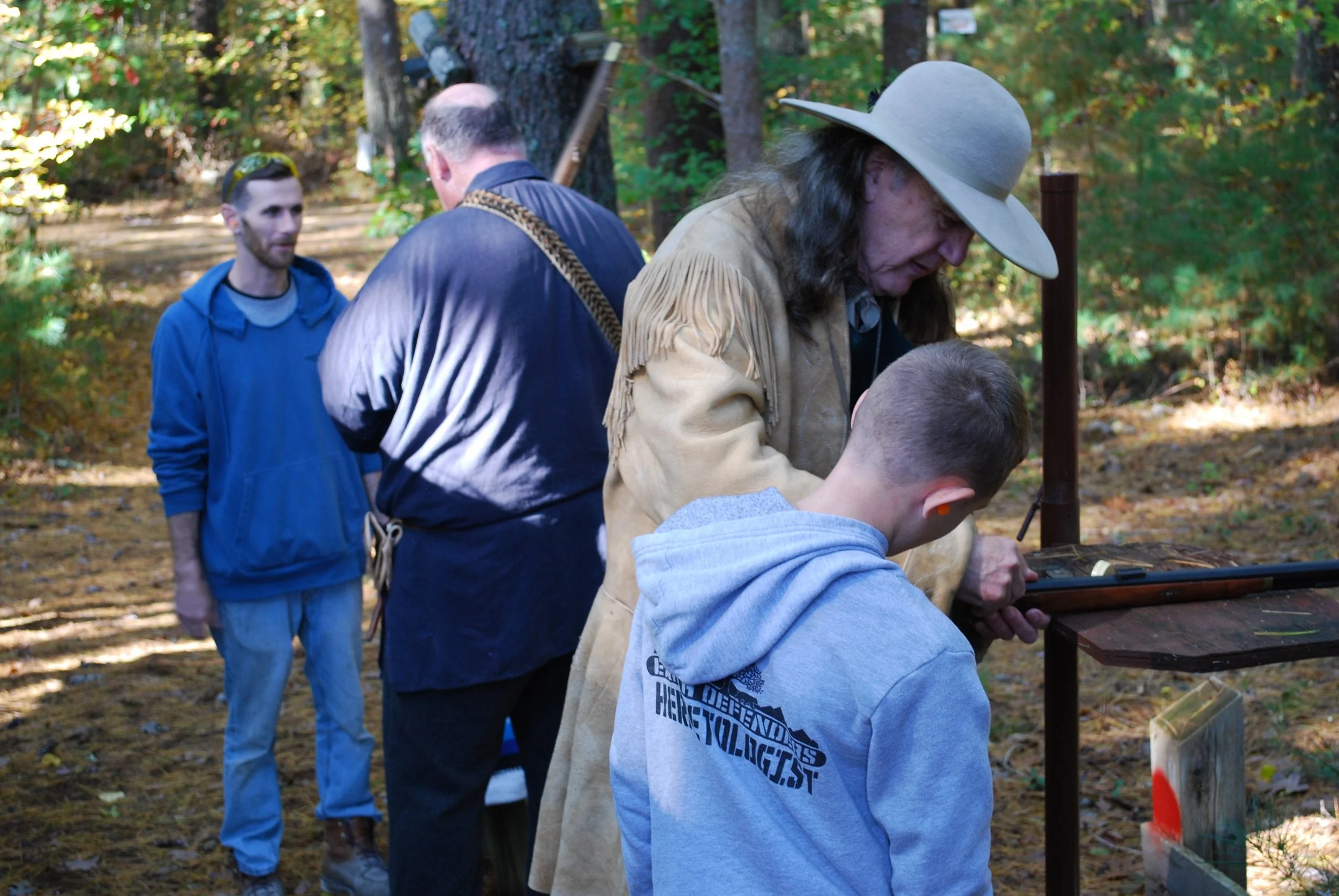 Four people are gathered outdoors in a wooded area. An older woman wearing a wide-brimmed hat and tan jacket appears to be showing something to a young boy in a gray hoodie. Two men, one in a blue hoodie and another with his back turned wearing a dar