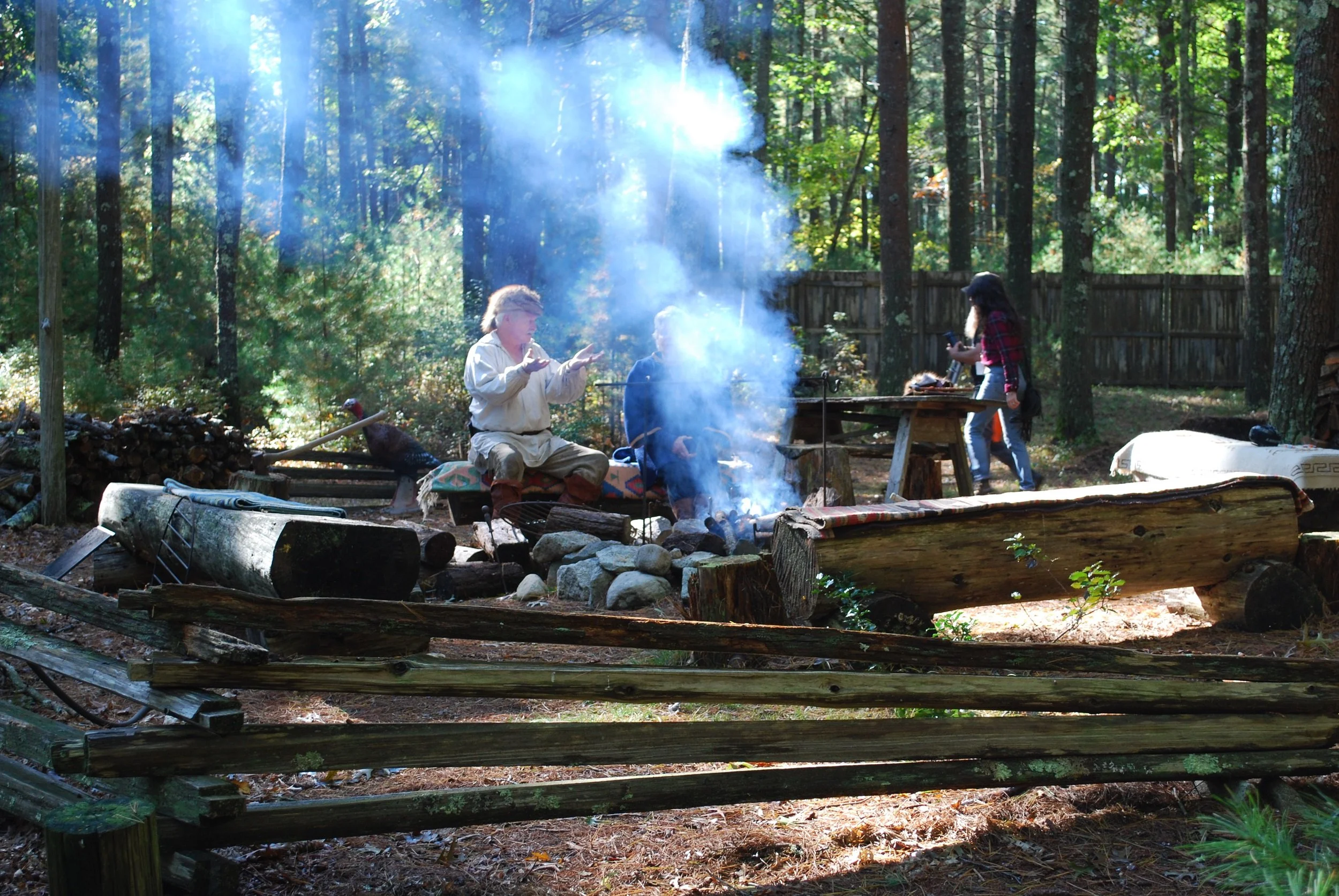 People sitting around a campfire in a forest, with a woman walking nearby, surrounded by trees and a wooden fence in the background.