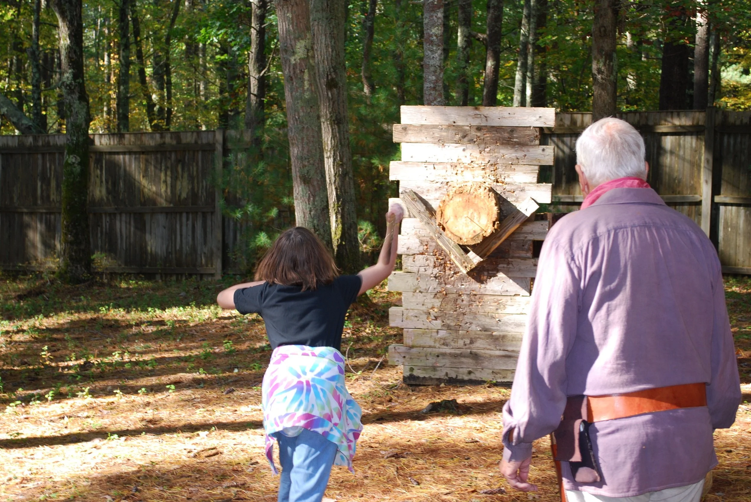 A young girl and an elderly man are outside in a wooded area. The girl is throwing a tomahawk into a stack of cut wood, which is set against a tree and a wooden fence. The elderly man is watching her.