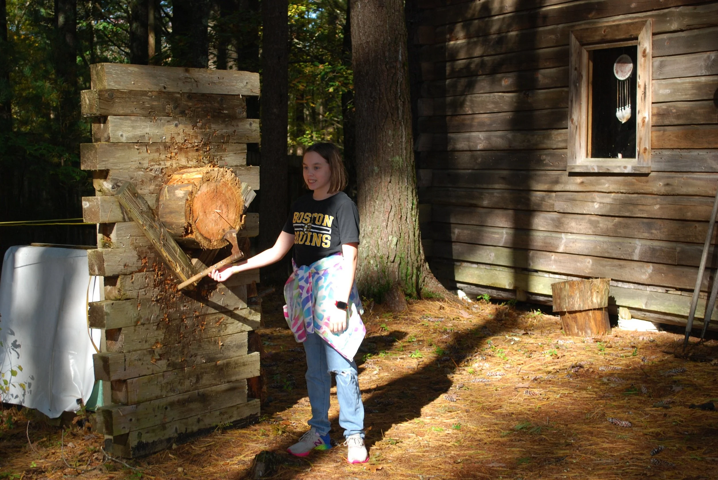 A girl in a Boston University T-shirt and colorful pants is throwing a tomahawk at a log near a wooden cabin in a forested area, with trees and sunlight in the background.