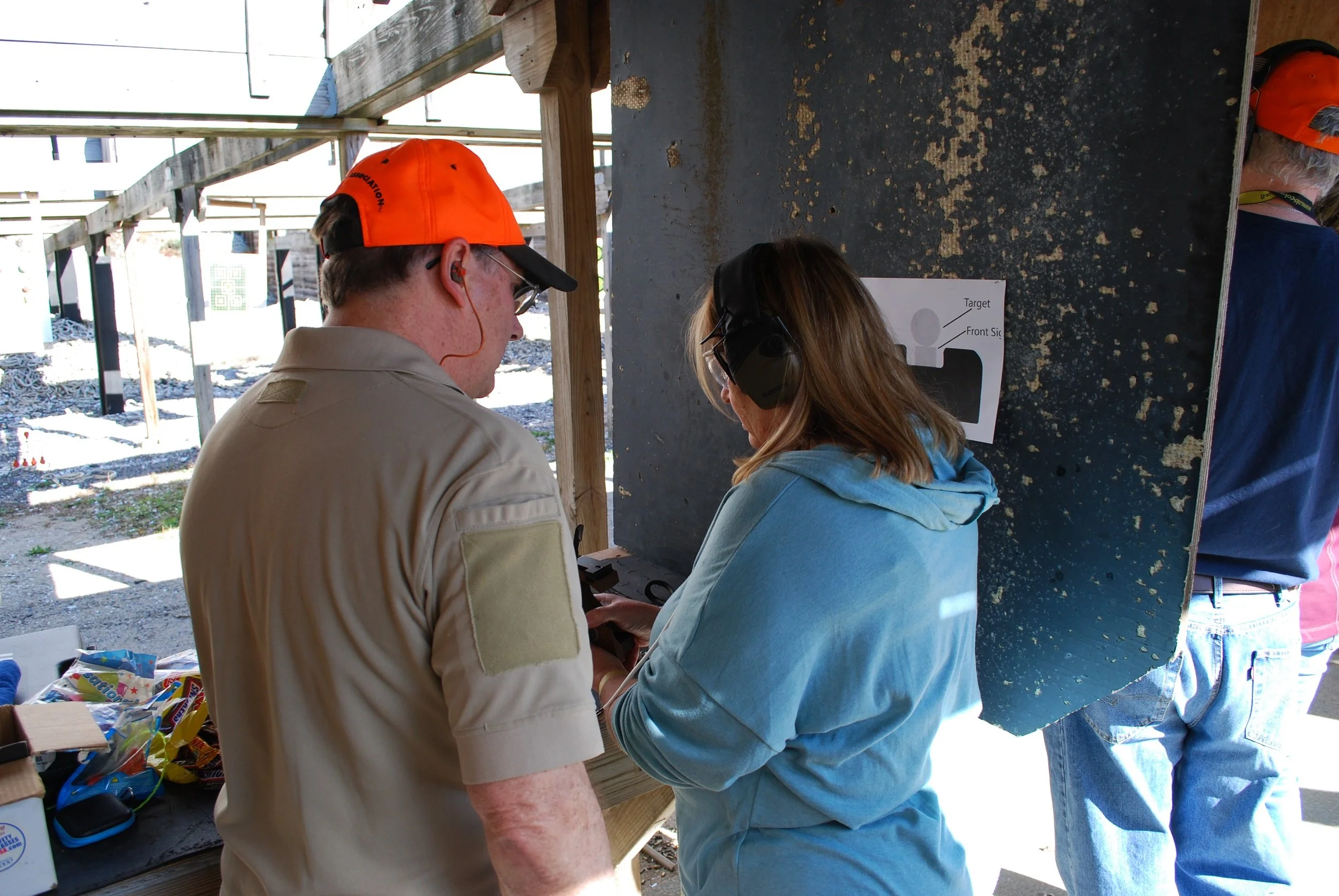 Two people, a man wearing orange cap and a woman with ear protection, at a shooting range. They are focusing on a target poster on the wall.