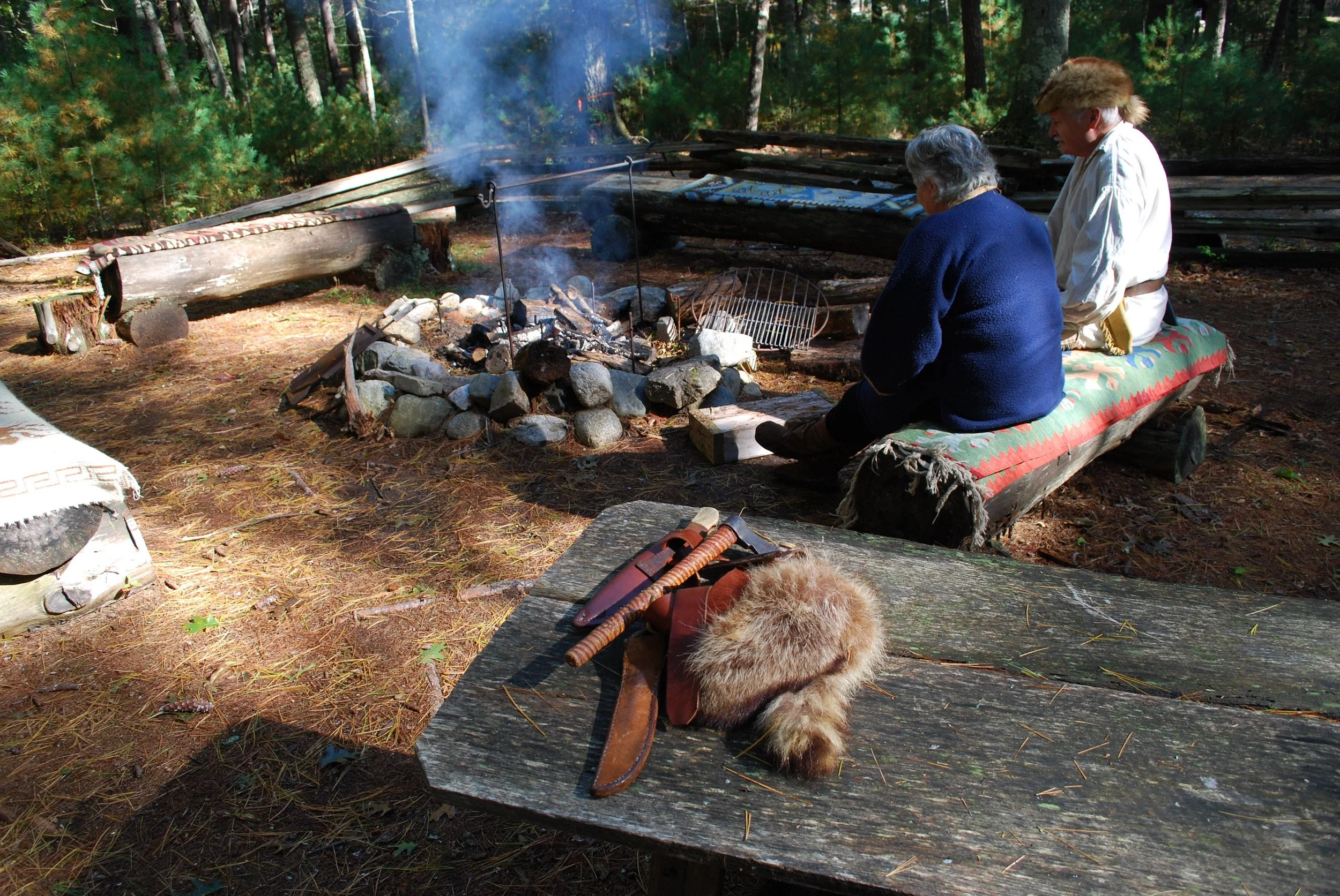Two sitting around a campfire in a forest, wearing traditional clothing. In the foreground, a wooden table has a fur hat, a knife, and a leather sheath. The campfire is surrounded by rocks and smoke rises from it. 