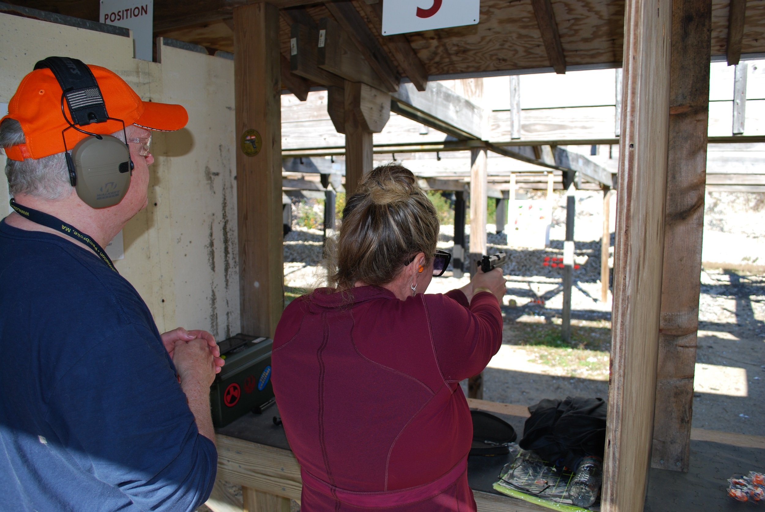 A woman in a maroon jacket aiming a handgun at a shooting range, with an older man in a blue shirt and orange cap watching her. They are in an outdoor shooting area with wooden structures.