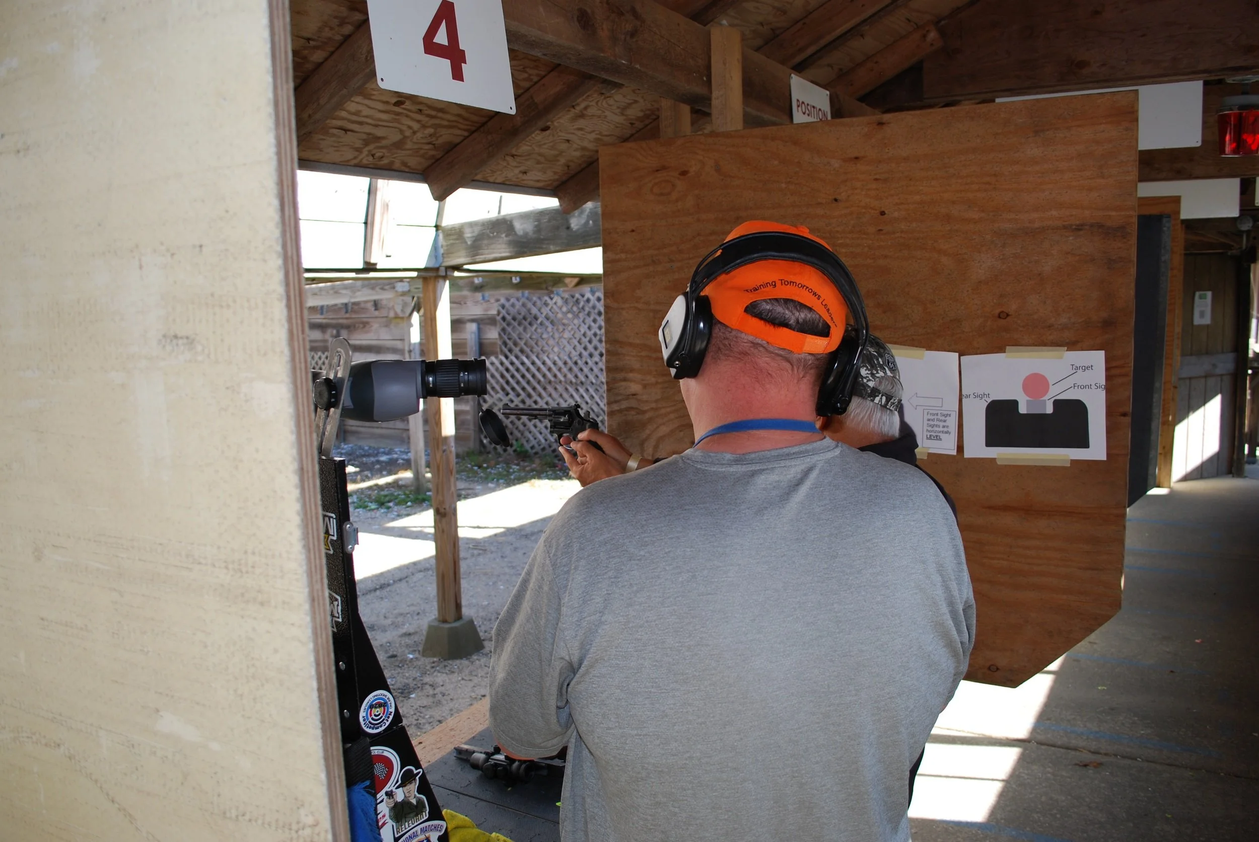 Man at outdoor shooting range wearing orange cap and headphones, aiming a pistol at a target.