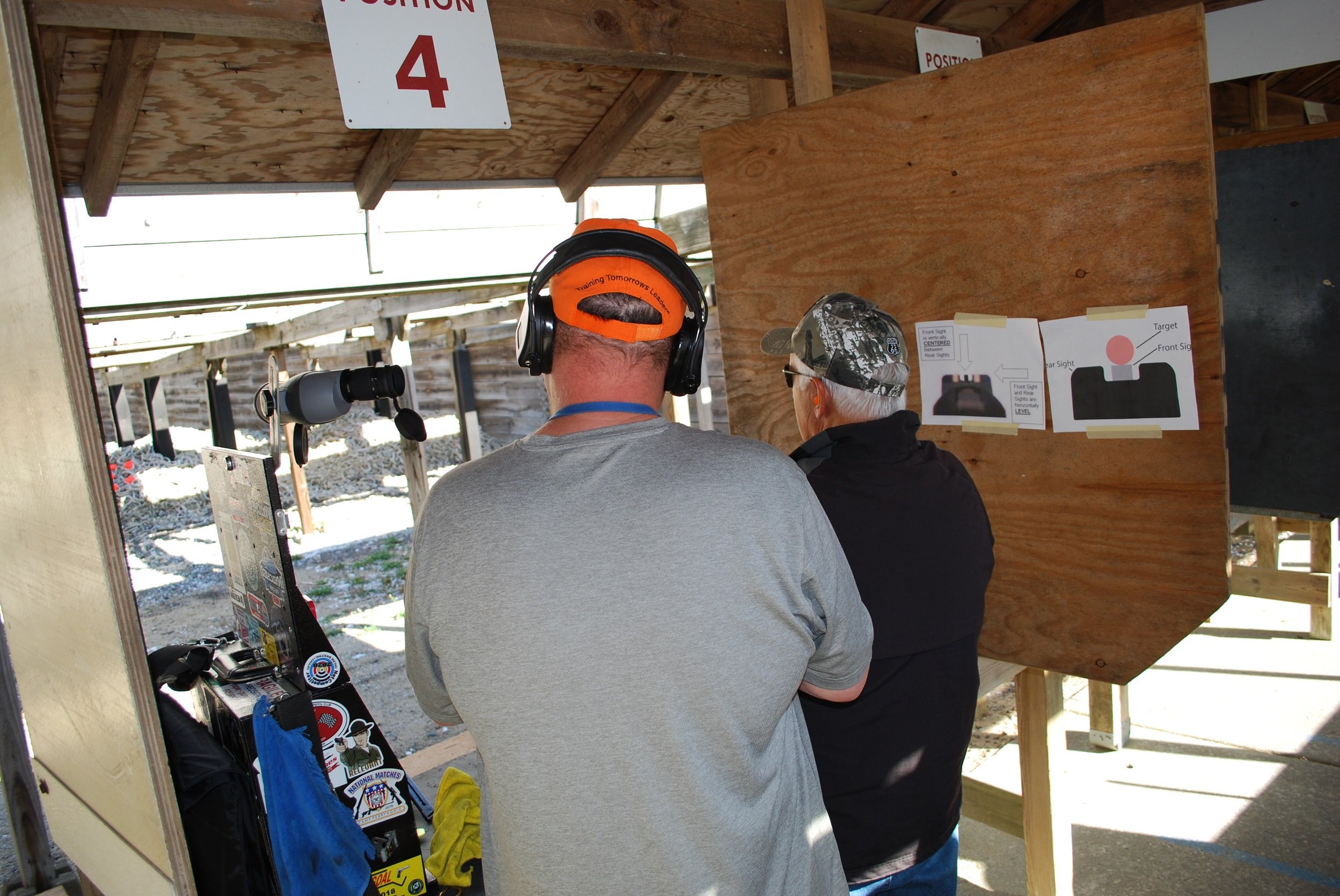 Two men at a shooting range, one with headphones and a gray shirt, the other with a camouflage cap and black jacket, standing in front of a target wall with diagram instructions.